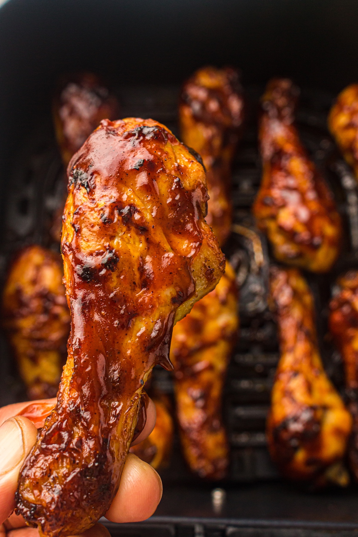 Close-up of a hand holding a glossy, barbecued chicken drumstick with caramelized sauce, against a blurred background of more drumsticks on a grill.