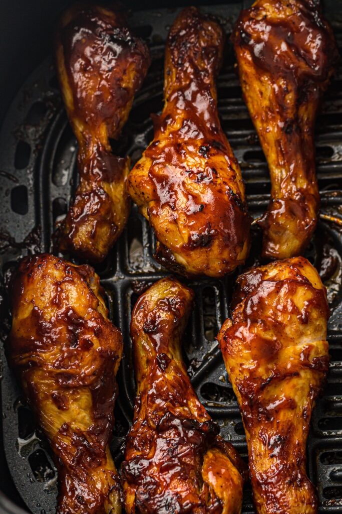 Close-up of barbecued chicken drumsticks coated in glossy, caramelized barbecue sauce, resting in an air fryer basket.