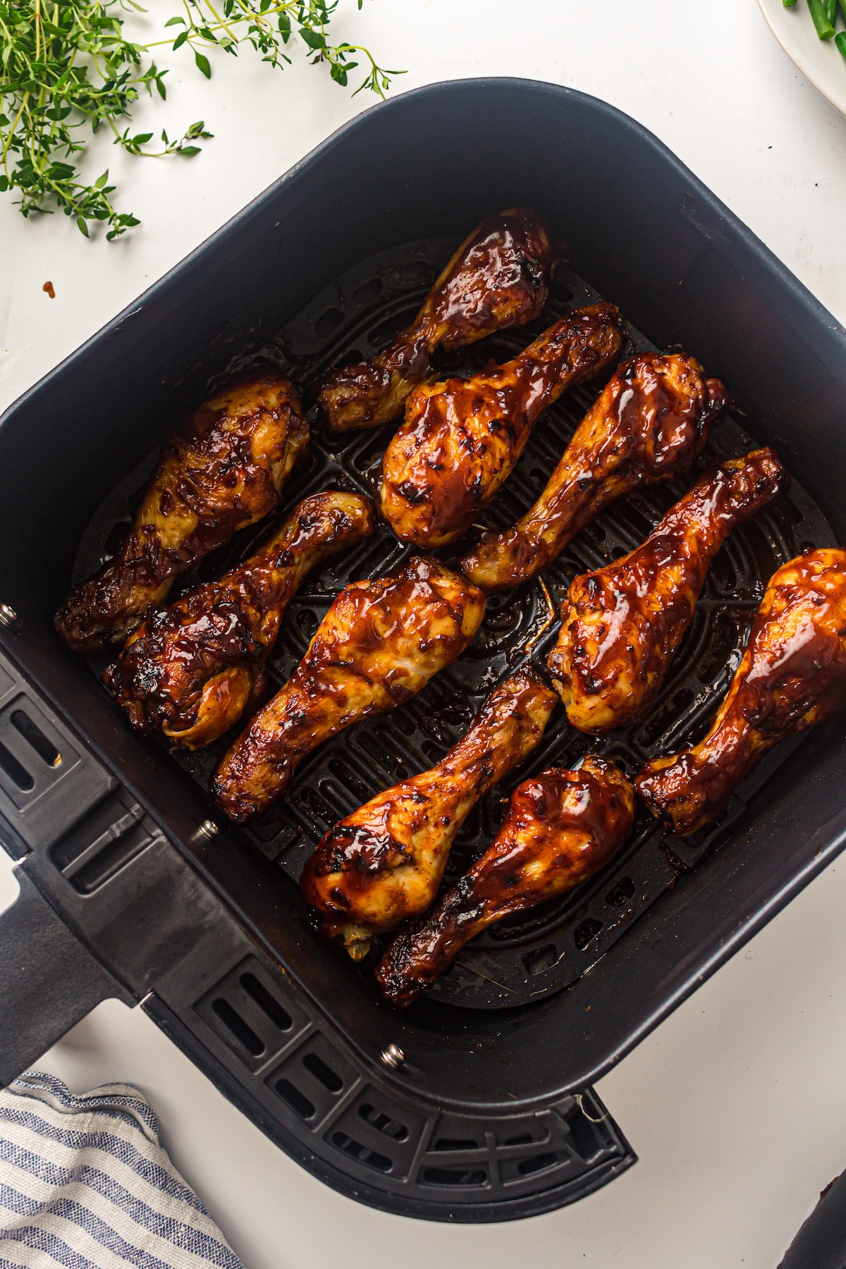Overhead view of barbecue-glazed chicken drumsticks in a black air fryer basket, with fresh herbs and a striped towel on a light surface.