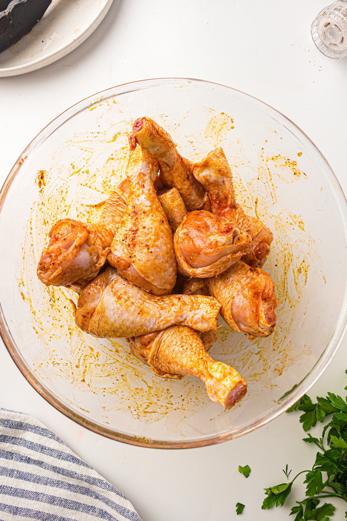 Top-down view of marinated chicken drumsticks in a glass bowl, surrounded by tongs, a spice grinder, parsley, and a striped towel on a light surface.