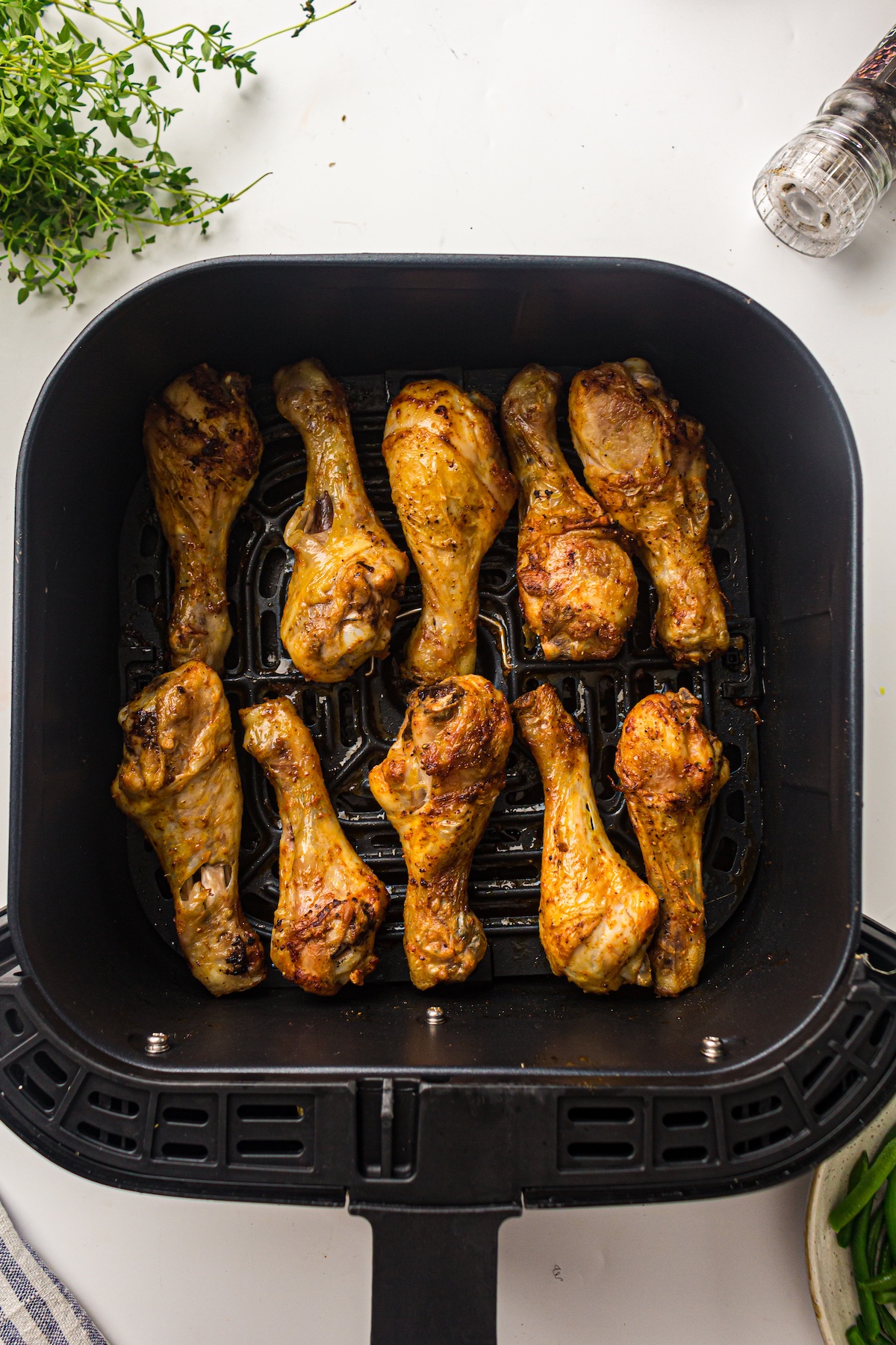 Overhead view of golden-brown chicken drumsticks in a black air fryer basket, surrounded by herbs, a spice grinder, a towel, and green beans.