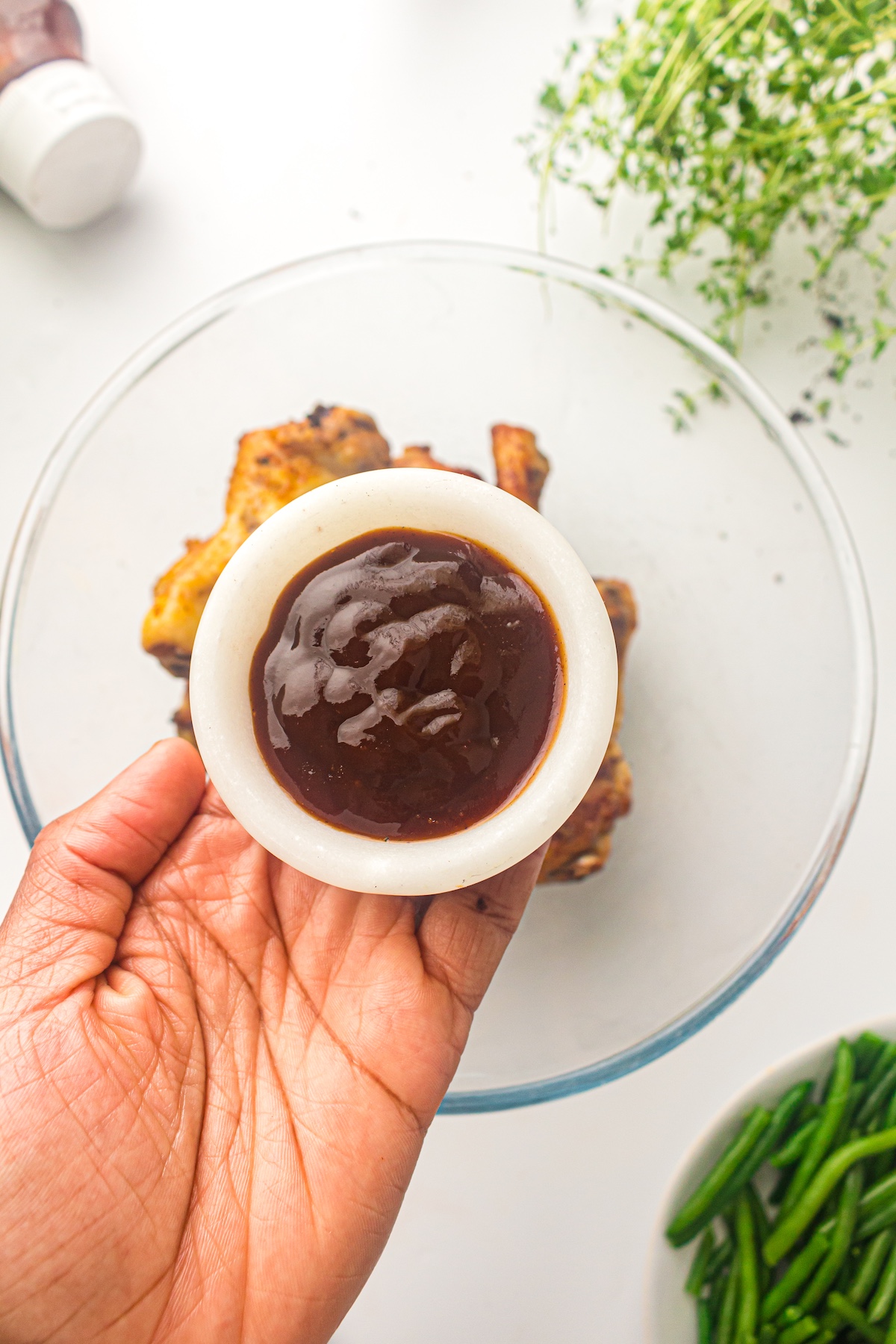 Top-down view of a hand holding a white bowl of dark brown sauce over a glass bowl of cooked chicken, with green beans and herbs nearby.