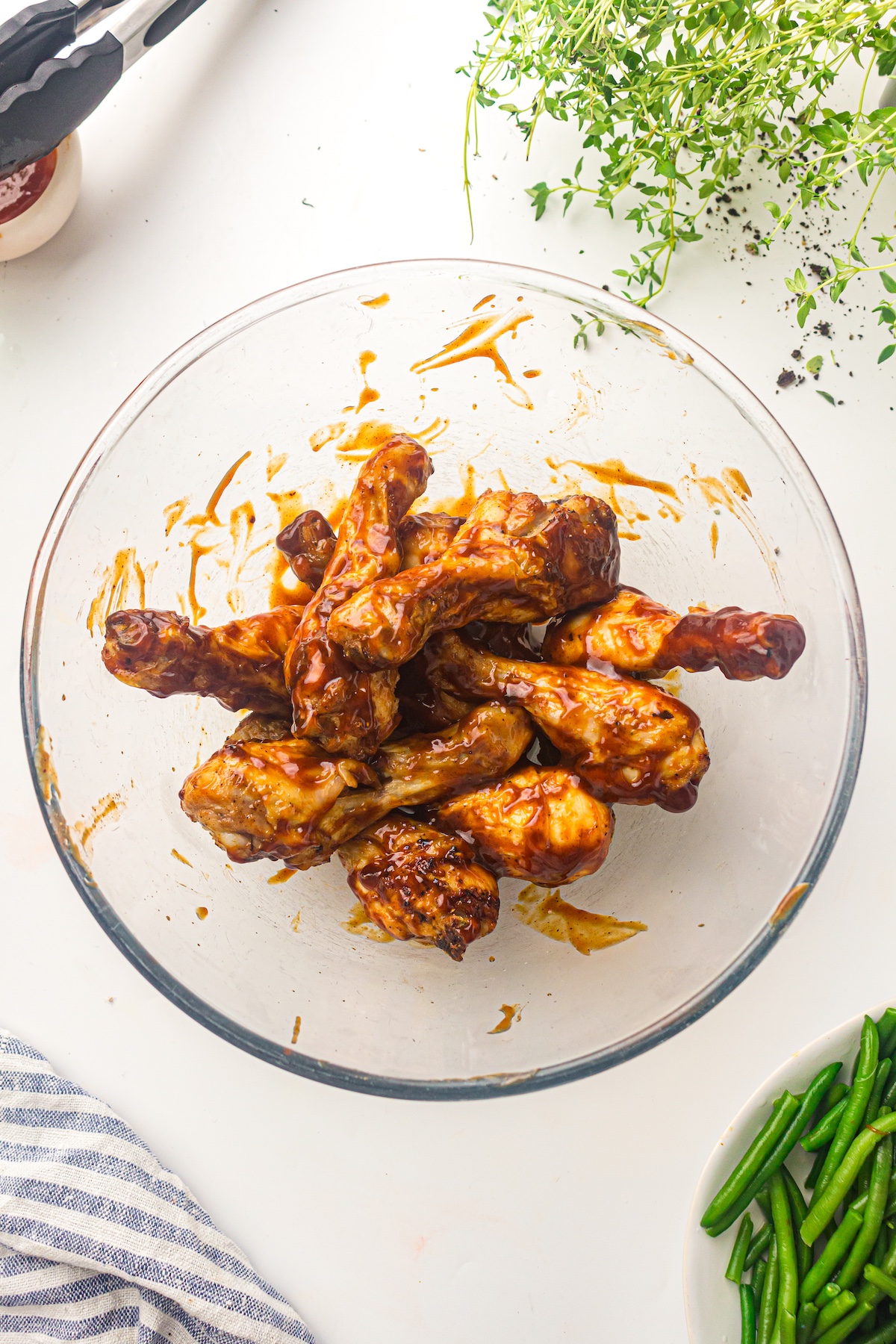 Overhead view of a glass bowl filled with barbecue-sauced chicken drumsticks, surrounded by tongs, herbs, a striped towel, and green beans.