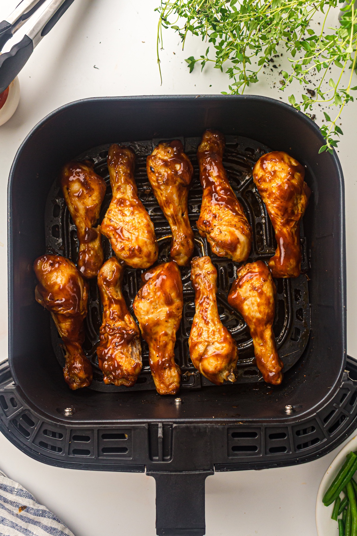 Overhead view of an air fryer basket with barbecue-sauced chicken drumsticks, surrounded by tongs, herbs, a striped towel, and green beans.