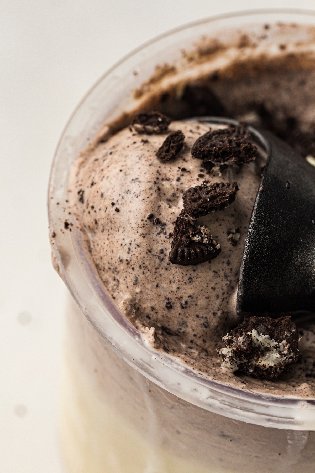 Close-up of cookies and cream ice cream in a clear plastic cup, topped with crushed chocolate cookie pieces, with a black spoon partially submerged.
