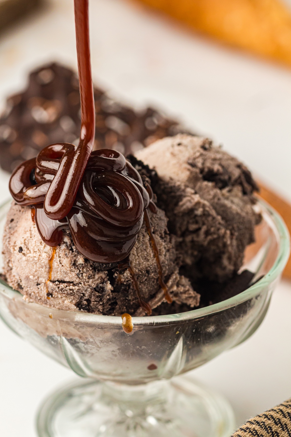 Close-up of two scoops of cookies and cream ice cream in a clear glass bowl, being drizzled with thick, glossy chocolate or caramel sauce.