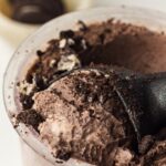 Close-up of cookies and cream ice cream in a clear plastic cup, topped with crushed chocolate cookie pieces, with a black spoon partially submerged.