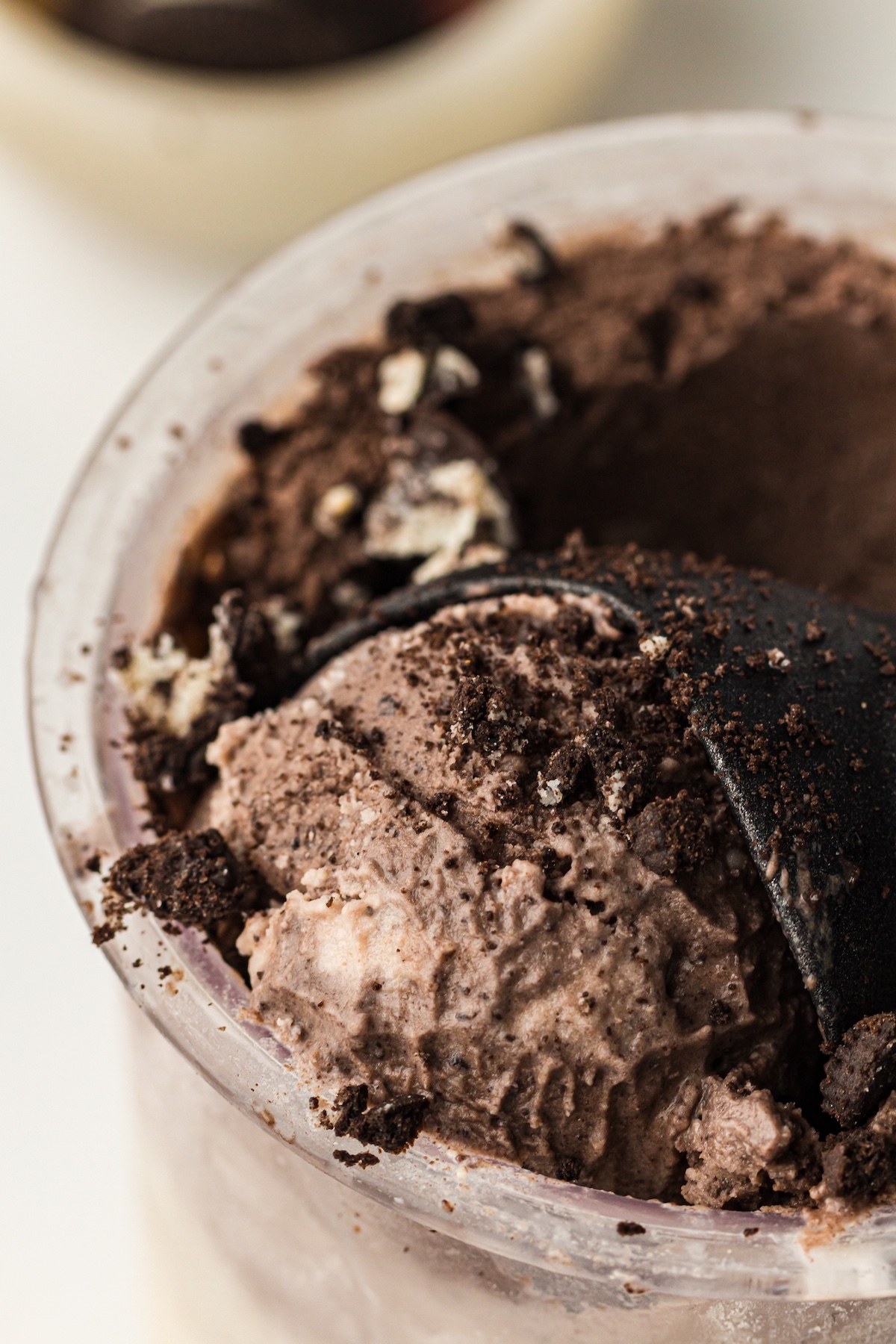 Close-up of cookies and cream ice cream in a clear plastic cup, topped with crushed chocolate cookie pieces, with a black spoon partially submerged.
