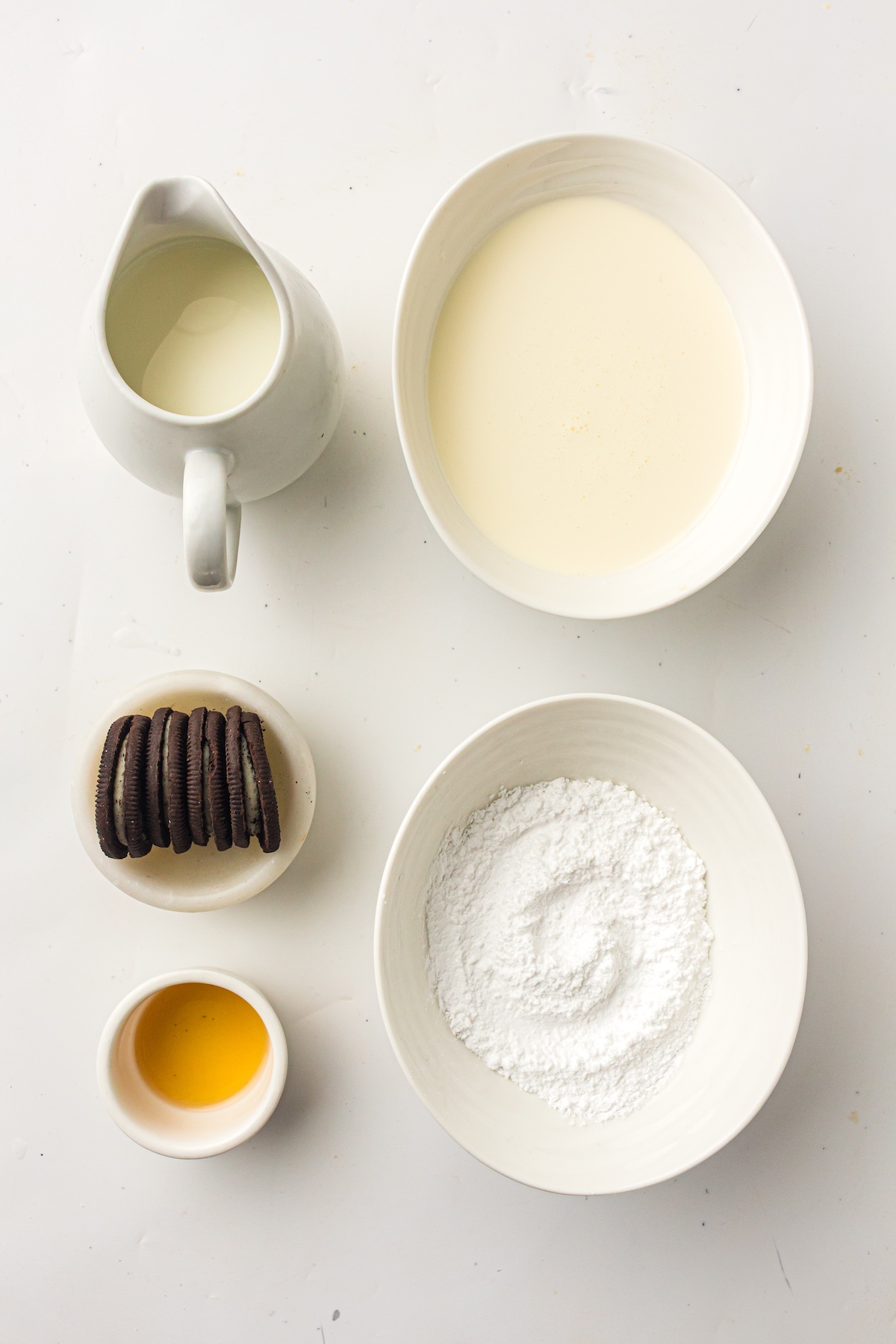 Overhead view of dessert ingredients on a white surface, including a creamer pitcher, Oreo cookies, powdered sugar, and vanilla extract.