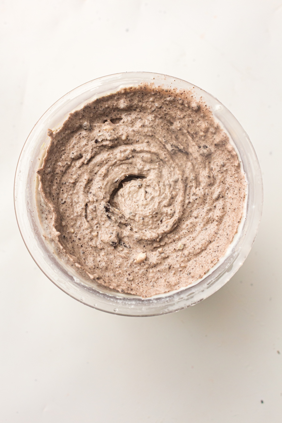 Close-up of a clear container filled with cookies and cream ice cream, showing a creamy, speckled texture with dark cookie crumbs and swirls.