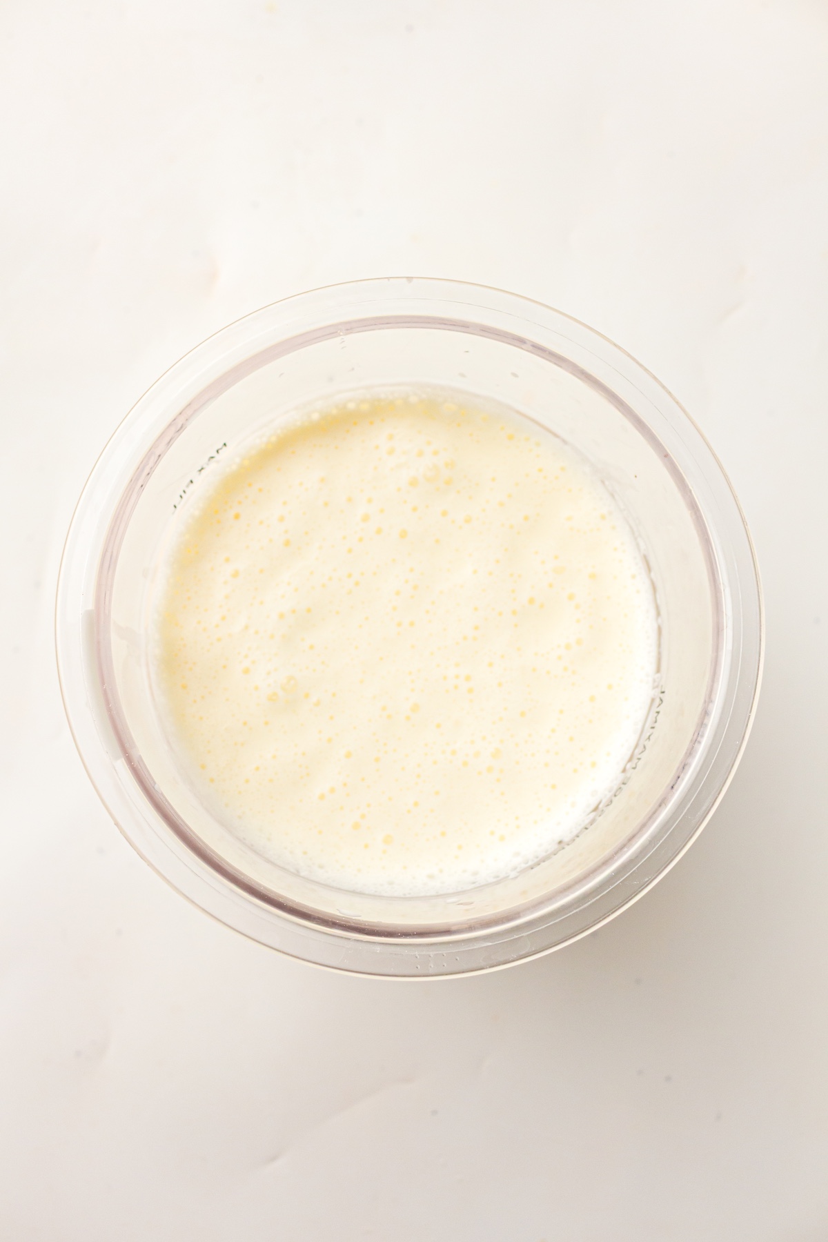 Top-down view of a clear glass bowl filled with pale yellow, frothy milk mixture, set on a light countertop.