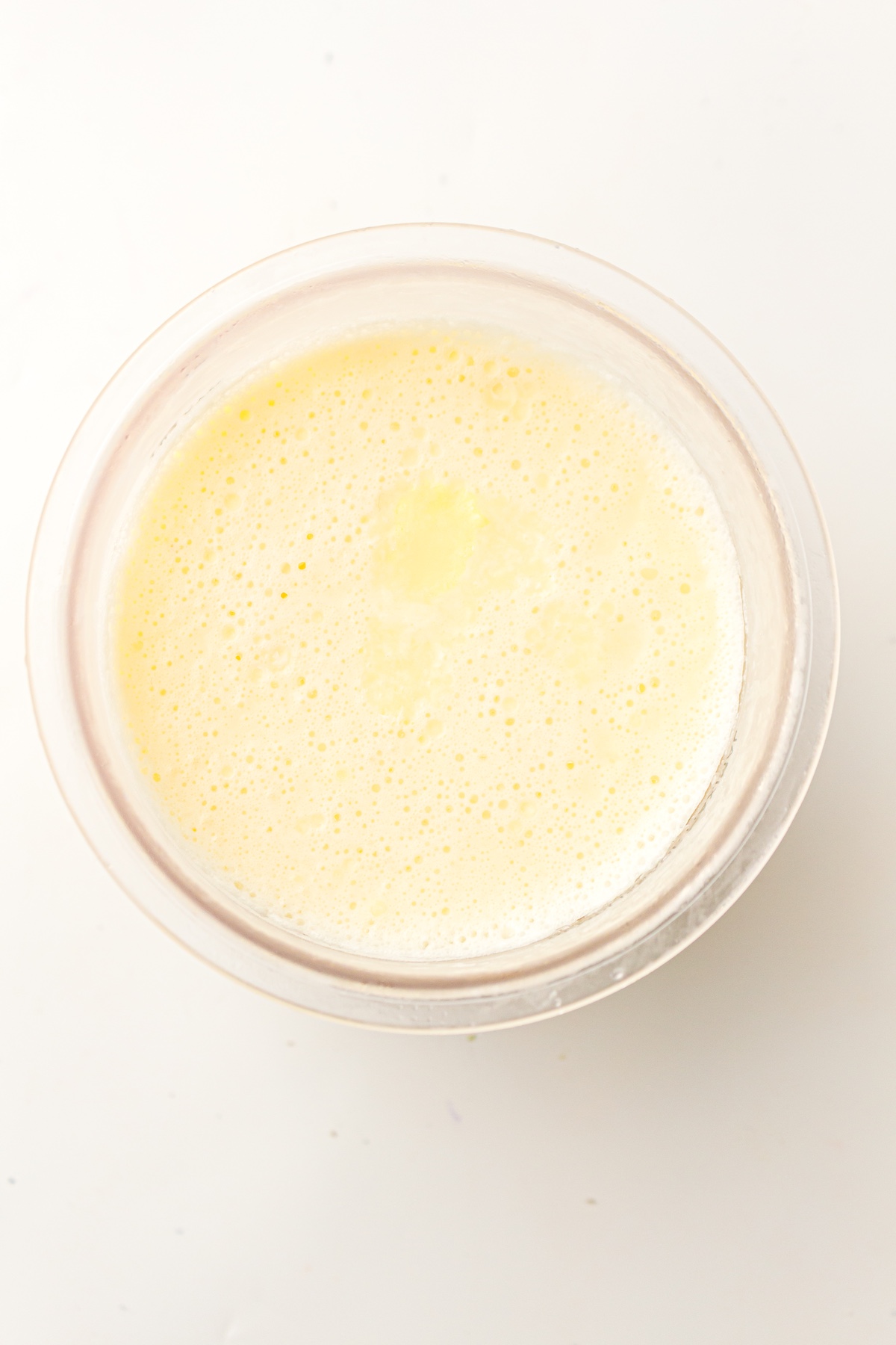 Top-down view of a clear bowl filled with a frozen, frothy, pale yellow milk mixture, set against a bright white background.
