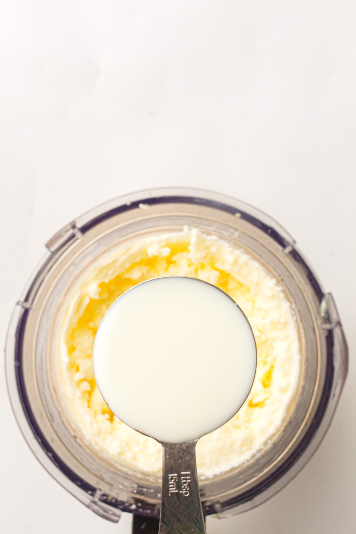 Overhead view of a silver tablespoon labeled '1 tbsp 15 mL,' filled with white liquid, held above a container of pale yellow creamy mixture.
