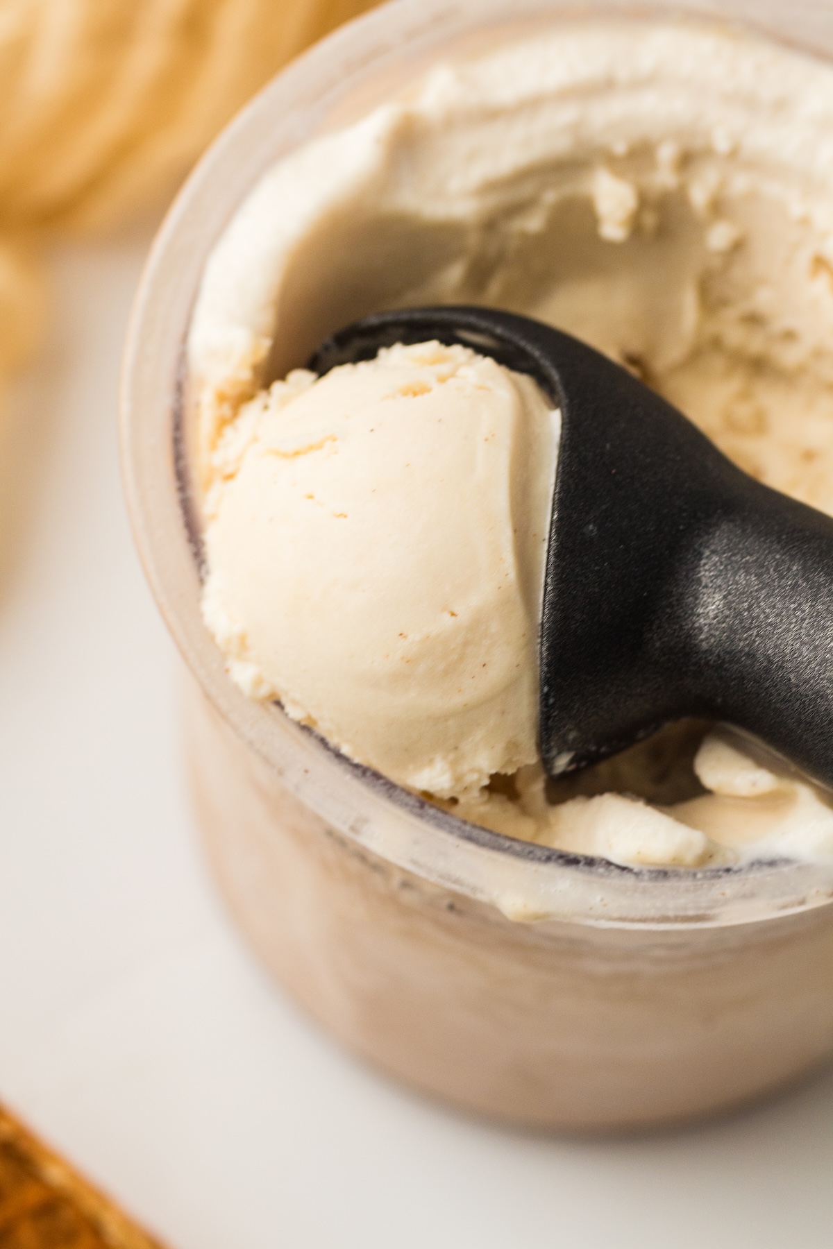 Close-up of a scoop of creamy, light-colored vanilla ice cream with dark specks, being lifted from a clear container by a black ice cream scoop, set against a softly blurred background with warm tones and a white surface.