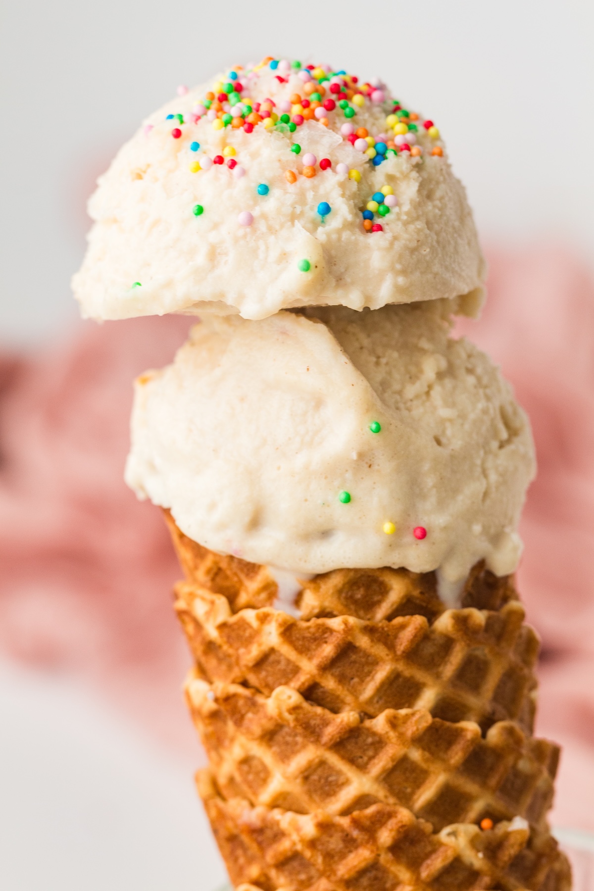 Close-up of two scoops of light-colored vanilla ice cream covered with colorful sprinkles, served in a golden-brown waffle cone, set against a blurred background with white and reddish-pink tones.