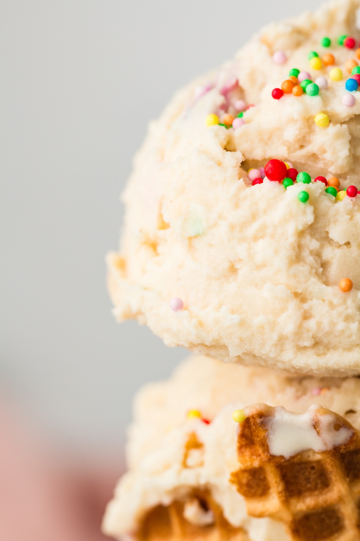 Close-up of a scoop of light-colored vanilla ice cream with colorful sprinkles, resting on a golden-brown waffle cone, set against a soft gray background.
