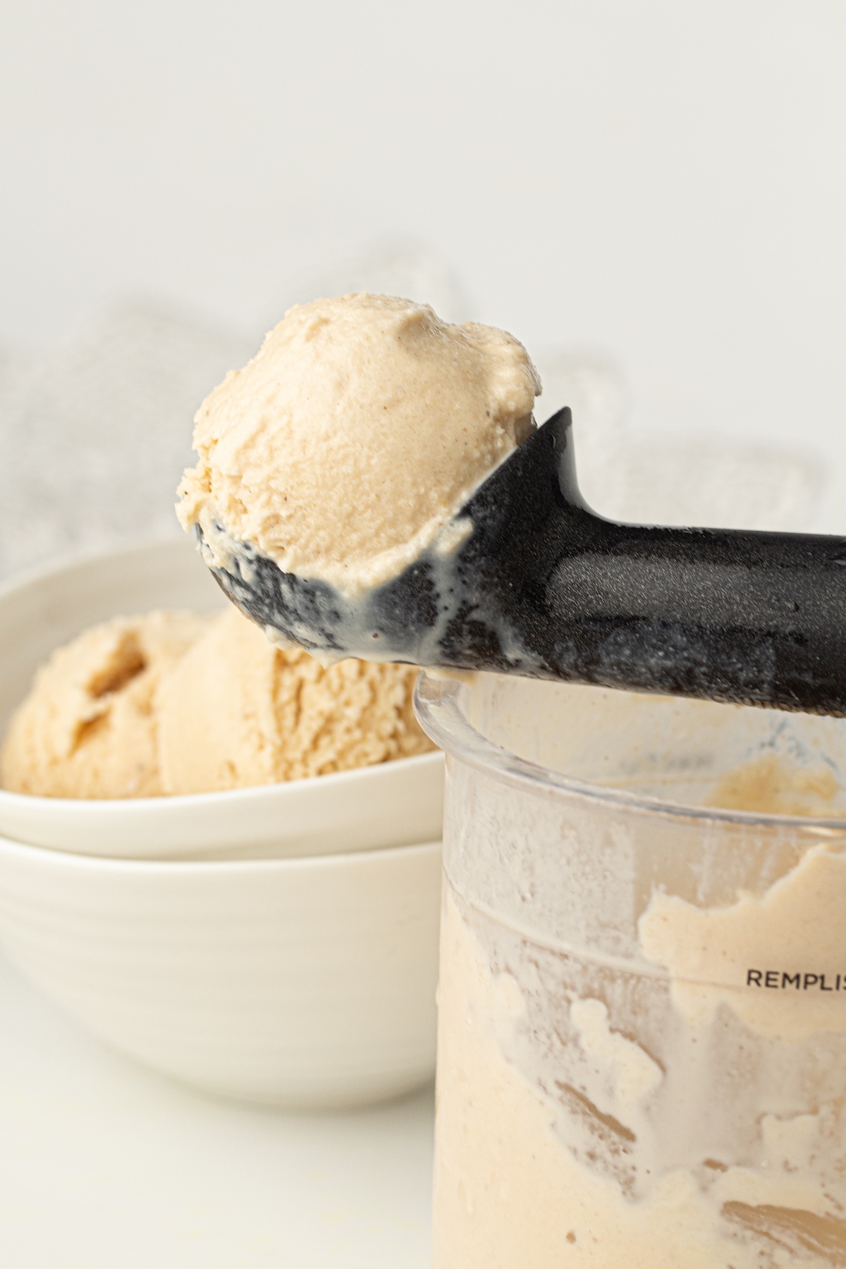 Close-up of a scoop of light-colored vanilla ice cream with dark specks, held by a black ice cream scoop above a clear container, with white bowls of ice cream visible in the blurred background.