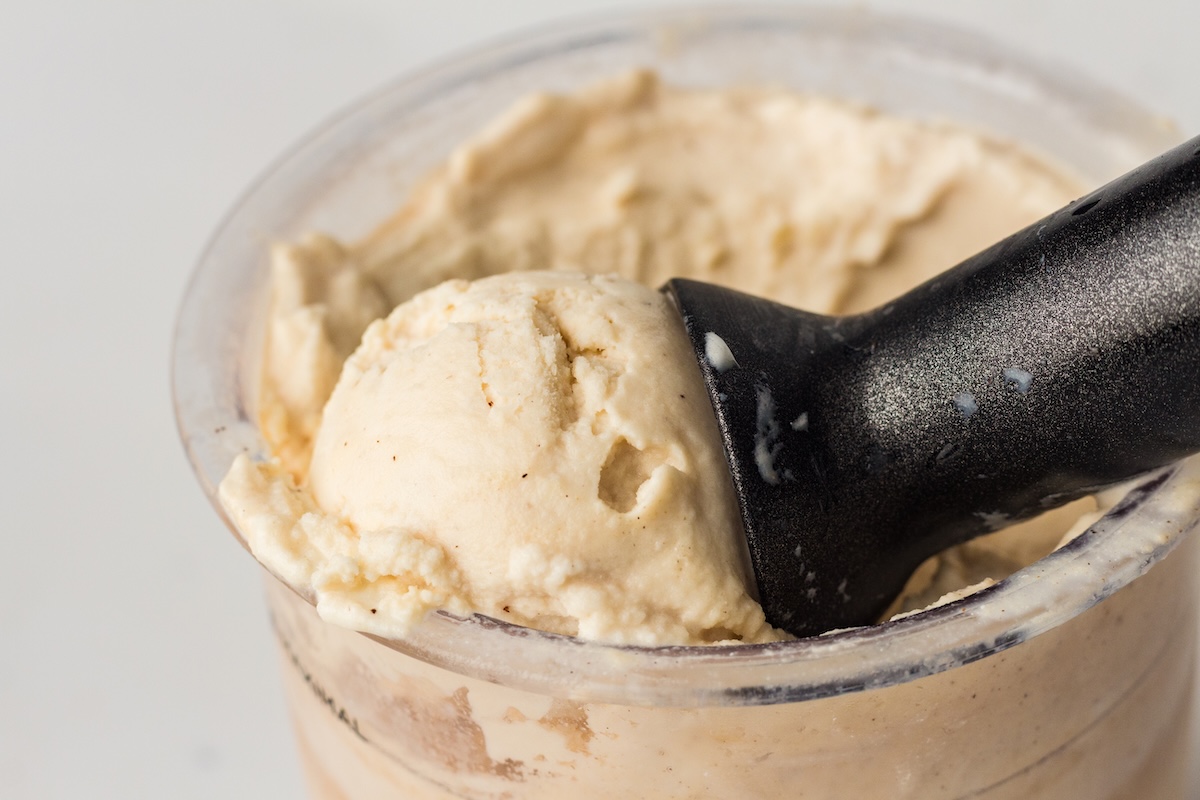 High-angle close-up of a black ice cream scoop lifting a rounded portion of light beige vanilla ice cream with dark specks from a clear container, set against a plain light background.