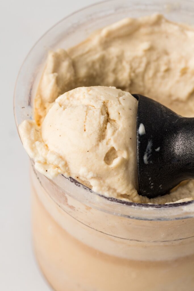 Close-up of a black ice cream scoop partially submerged in a clear container of light beige vanilla ice cream with dark specks, highlighting the creamy texture, set against a white background.