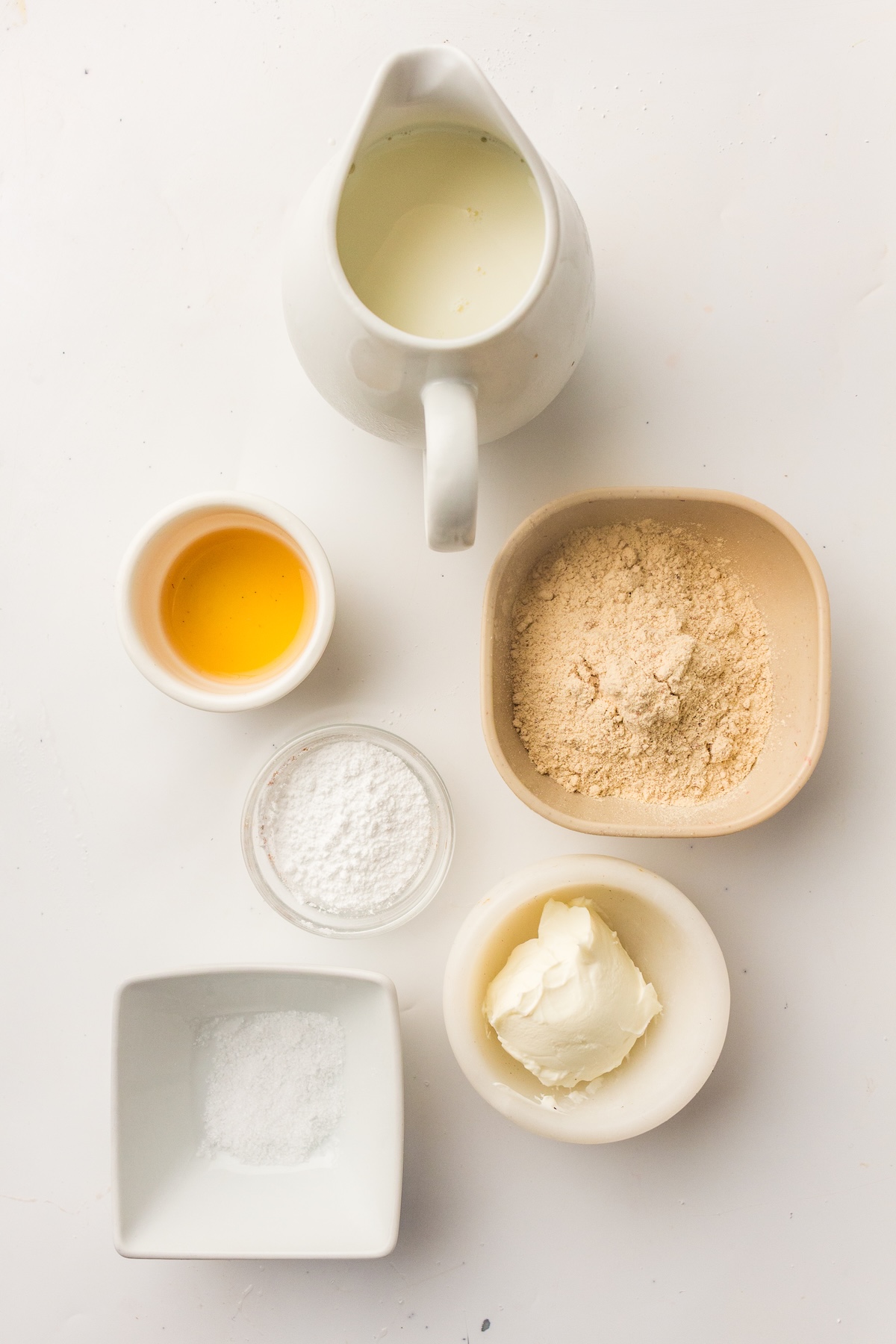 Overhead view of six ingredients for high-protein vanilla ice cream, including milk in a white pitcher, golden liquid in a small bowl, powdered sugar in a clear bowl, light brown powder in a beige bowl, white granules in a square bowl, and cream cheese in a round bowl, arranged on a white surface.