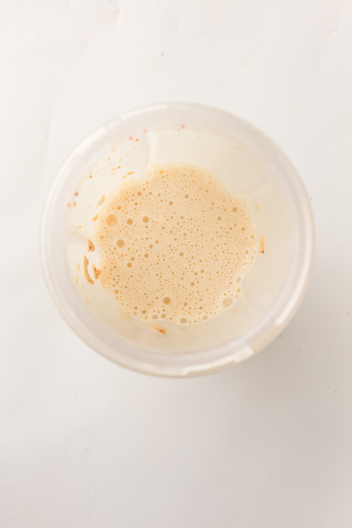 Overhead view of a clear cup containing light brown foamy liquid with small bubbles and darker specks on the inner walls, set against a clean white background.