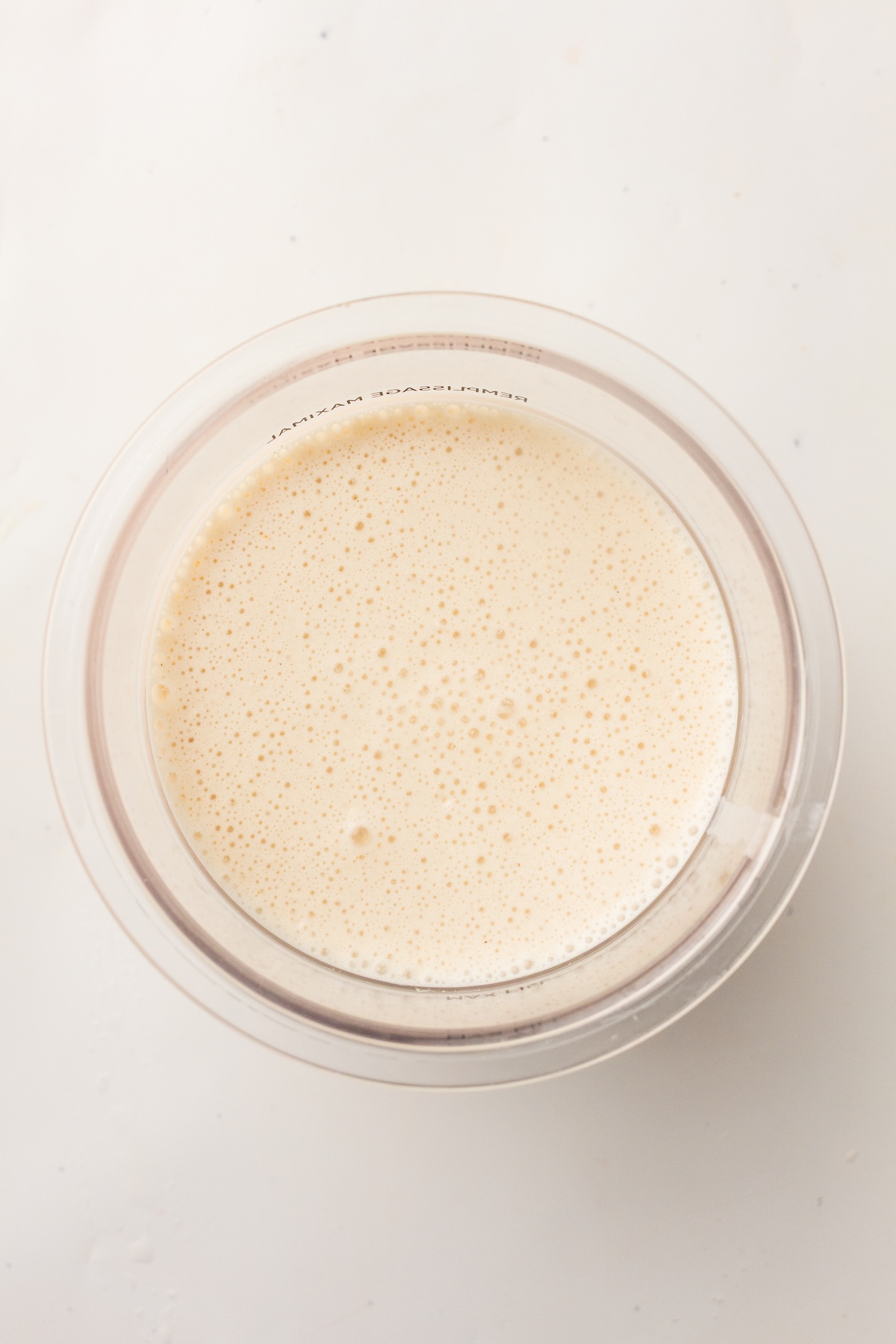 Top-down view of a clear container filled with light beige foamy liquid, with small bubbles covering the surface, and mirrored text on the container indicating 'Maximum Fill' in French, set against a light background.
