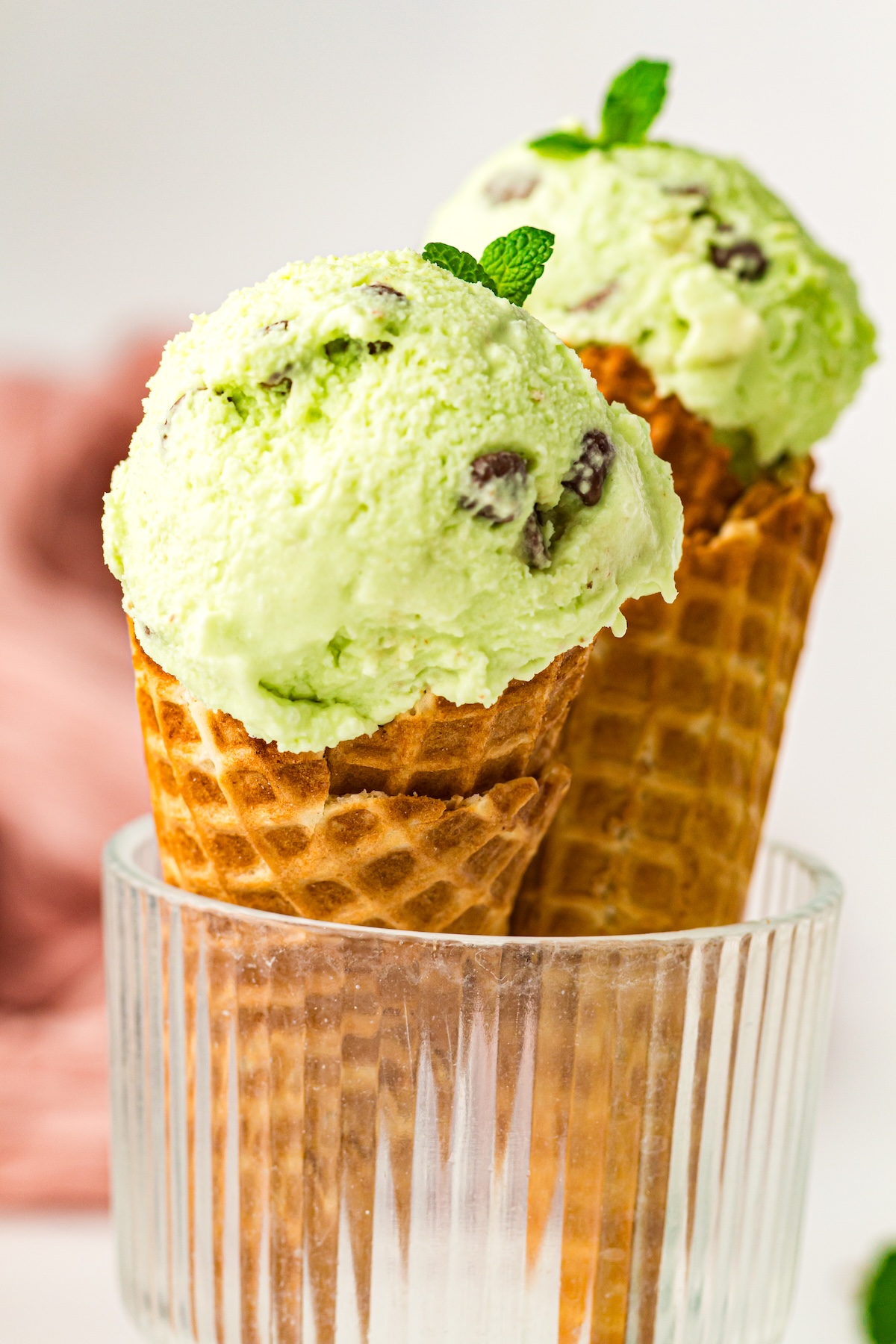 Close-up of two mint chocolate chip ice cream cones in a clear ribbed glass, garnished with fresh mint leaves, set against a clean white background with a hint of soft pink fabric in the distance.