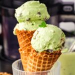 Two waffle cones filled with light green mint chocolate chip ice cream, garnished with fresh mint leaves, placed in a clear ribbed glass, with a softly blurred background featuring a dark appliance and light green tones.