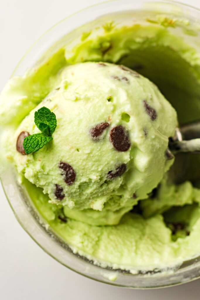 Overhead close-up of a scoop of light green mint chocolate chip ice cream, garnished with fresh mint leaves, being lifted from a clear glass container by a metal ice cream scoop, set against a bright white background.