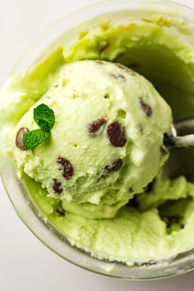 Overhead close-up of a scoop of light green mint chocolate chip ice cream, garnished with fresh mint leaves, being lifted from a clear glass container by a metal ice cream scoop, set against a bright white background.