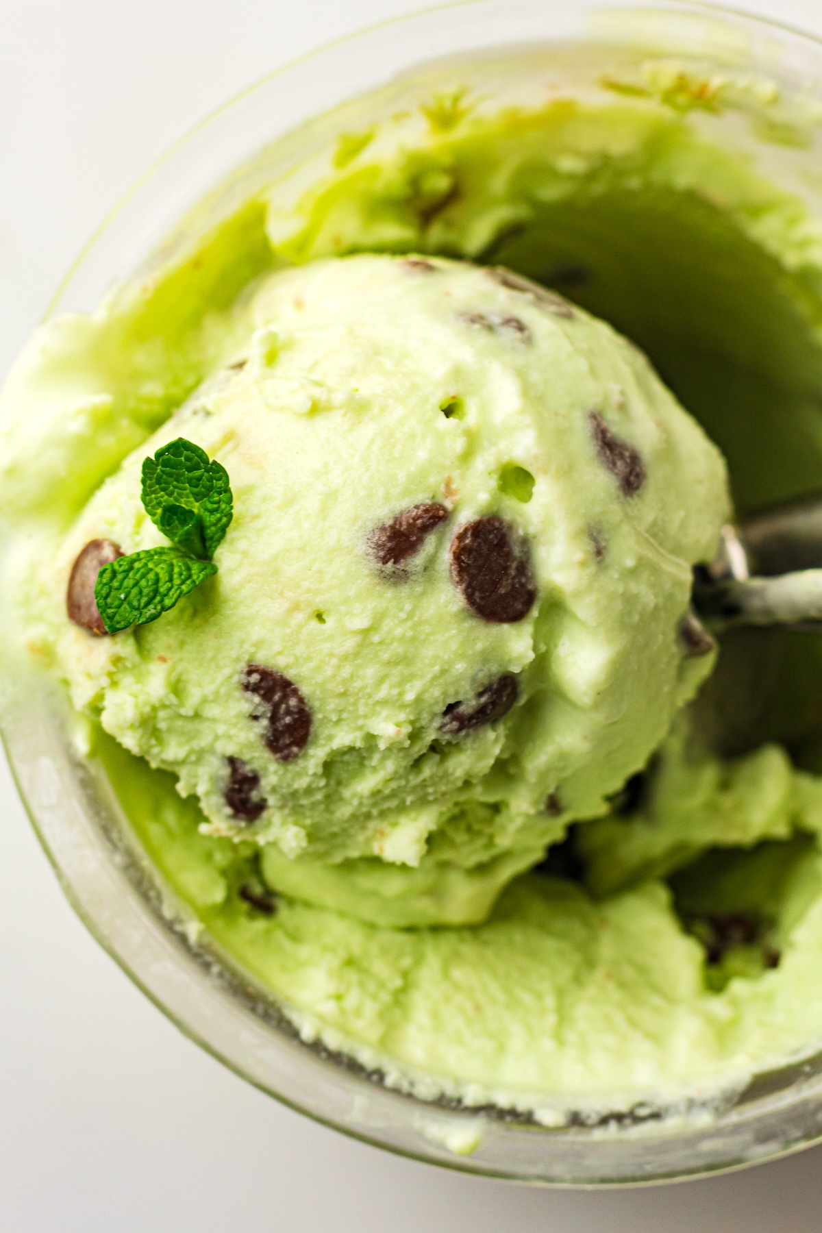 Overhead close-up of a scoop of light green mint chocolate chip ice cream, garnished with fresh mint leaves, being lifted from a clear glass container by a metal ice cream scoop, set against a bright white background.