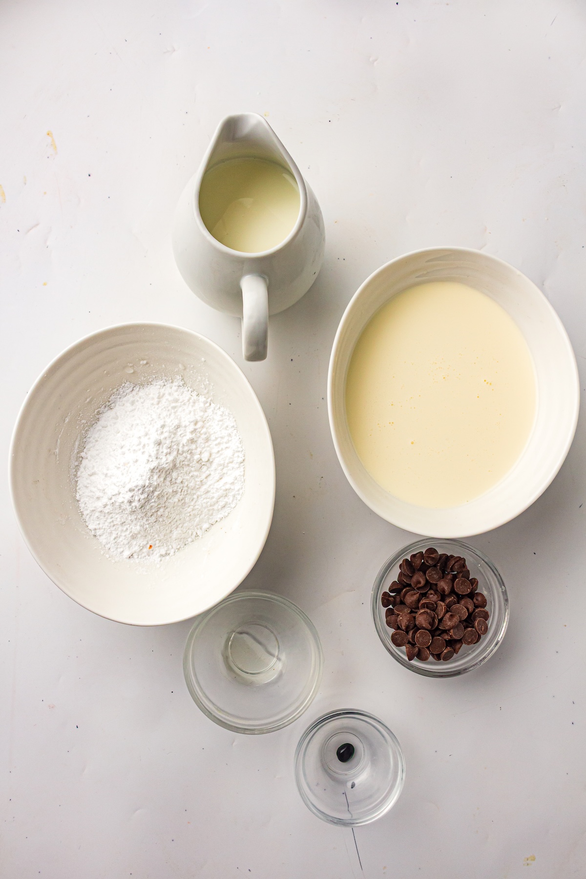 Flat lay of mint chocolate chip ice cream ingredients on a light surface, including a pitcher of cream, a bowl of powdered sugar, a bowl of chocolate chips, and an empty glass bowl.