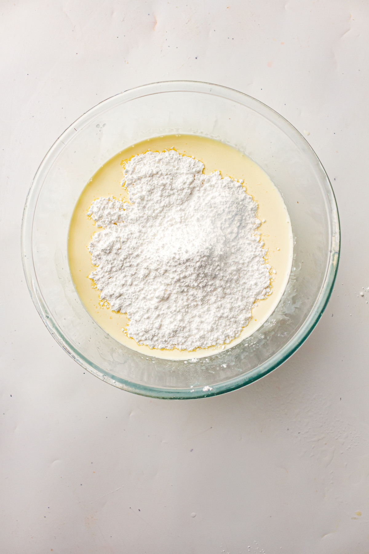 Overhead view of a clear glass bowl containing a light yellow liquid topped with a mound of white powdered sugar, set on a light-colored surface.