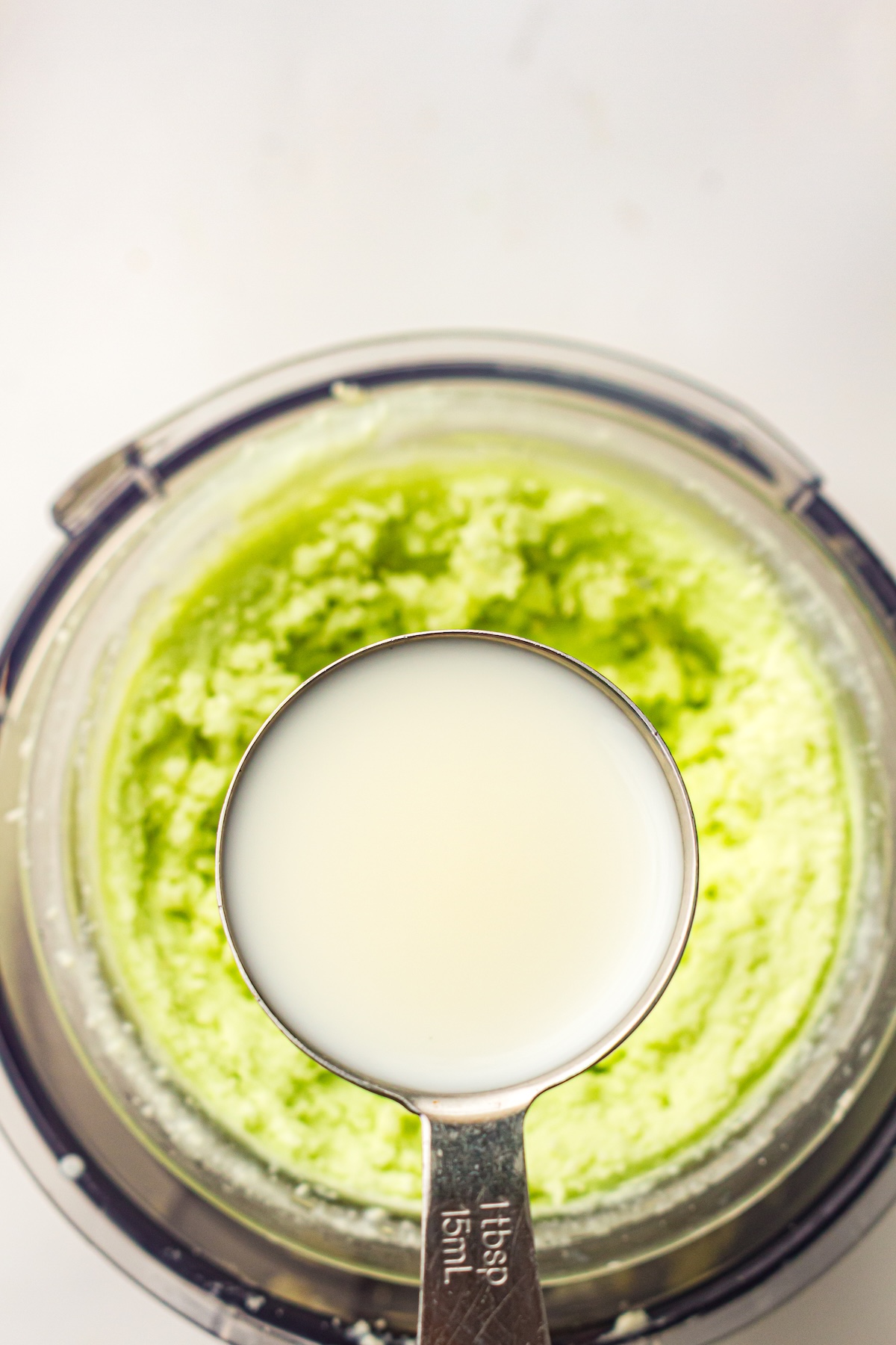 Top-down view of a clear bowl containing a vibrant green textured mixture, with a silver tablespoon labeled '1tbsp 15mL' held above, filled with a pale liquid, likely milk or cream, set against a clean white background.