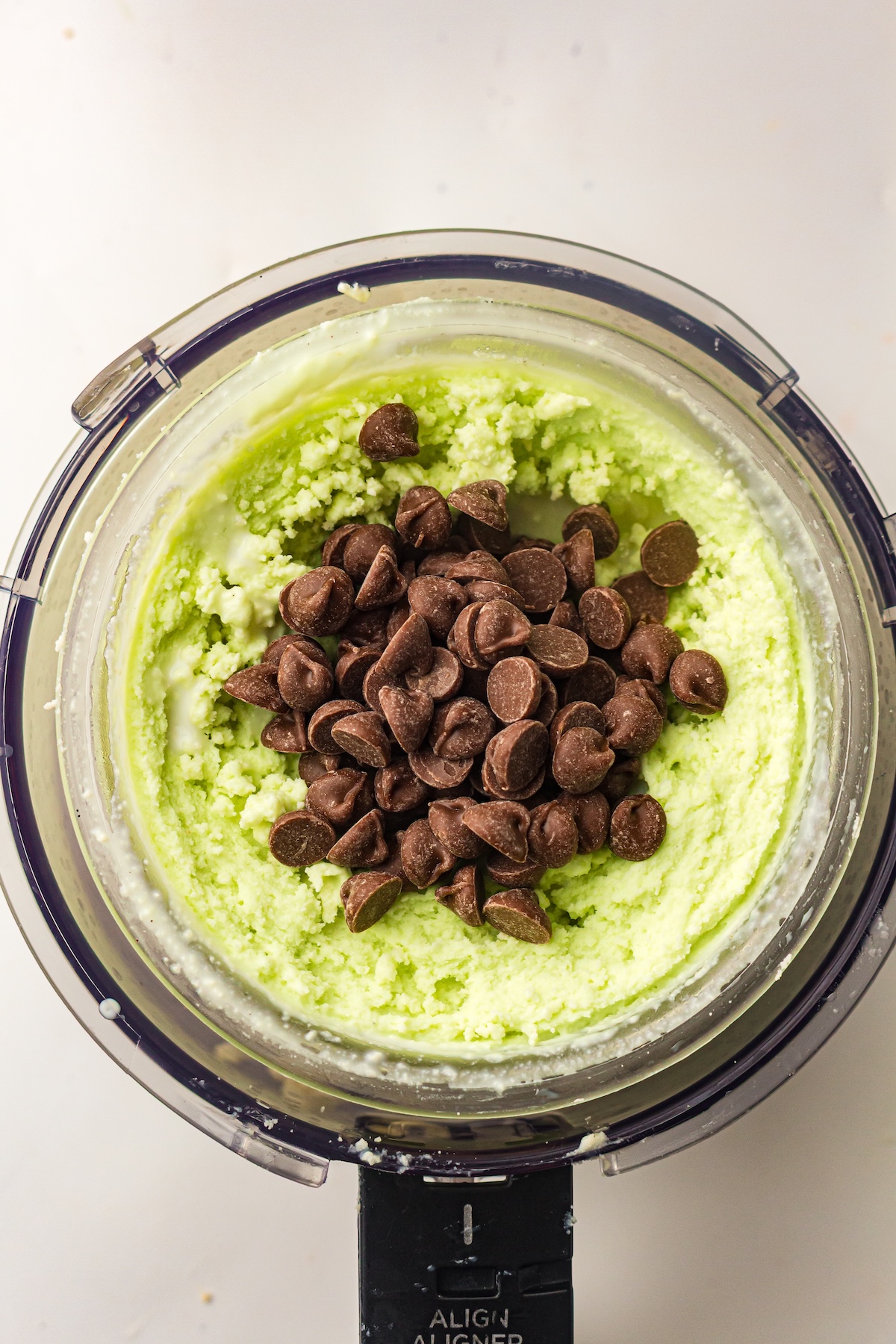 Overhead view of a food processor bowl filled with a light green, granular mixture topped with a mound of dark chocolate chips, with the appliance base visible below, labeled 'ALIGN ALIGNER,' set on a light background.