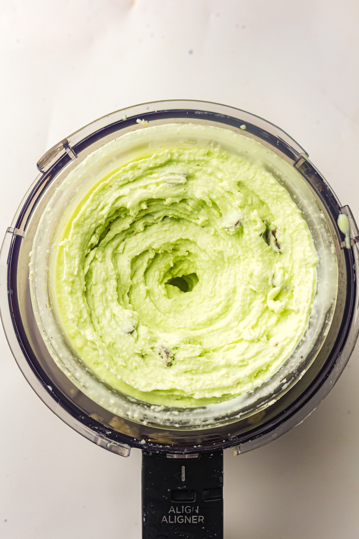 Top-down view of a clear food processor bowl containing a light green creamy mixture with swirls and darker specks, sitting on a black appliance base labeled 'ALIGN ALIGNER,' set against a light background.