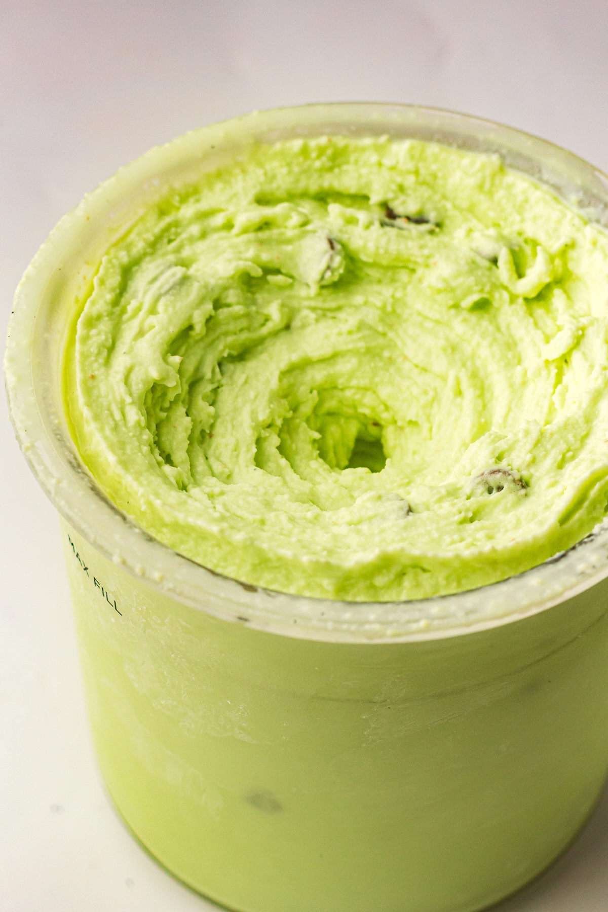 Close-up, top-down view of a light green creamy mixture with swirled textures and small dark inclusions, contained in a translucent cylindrical container with a 'MAX FILL' line visible on the side, set against a soft light background.