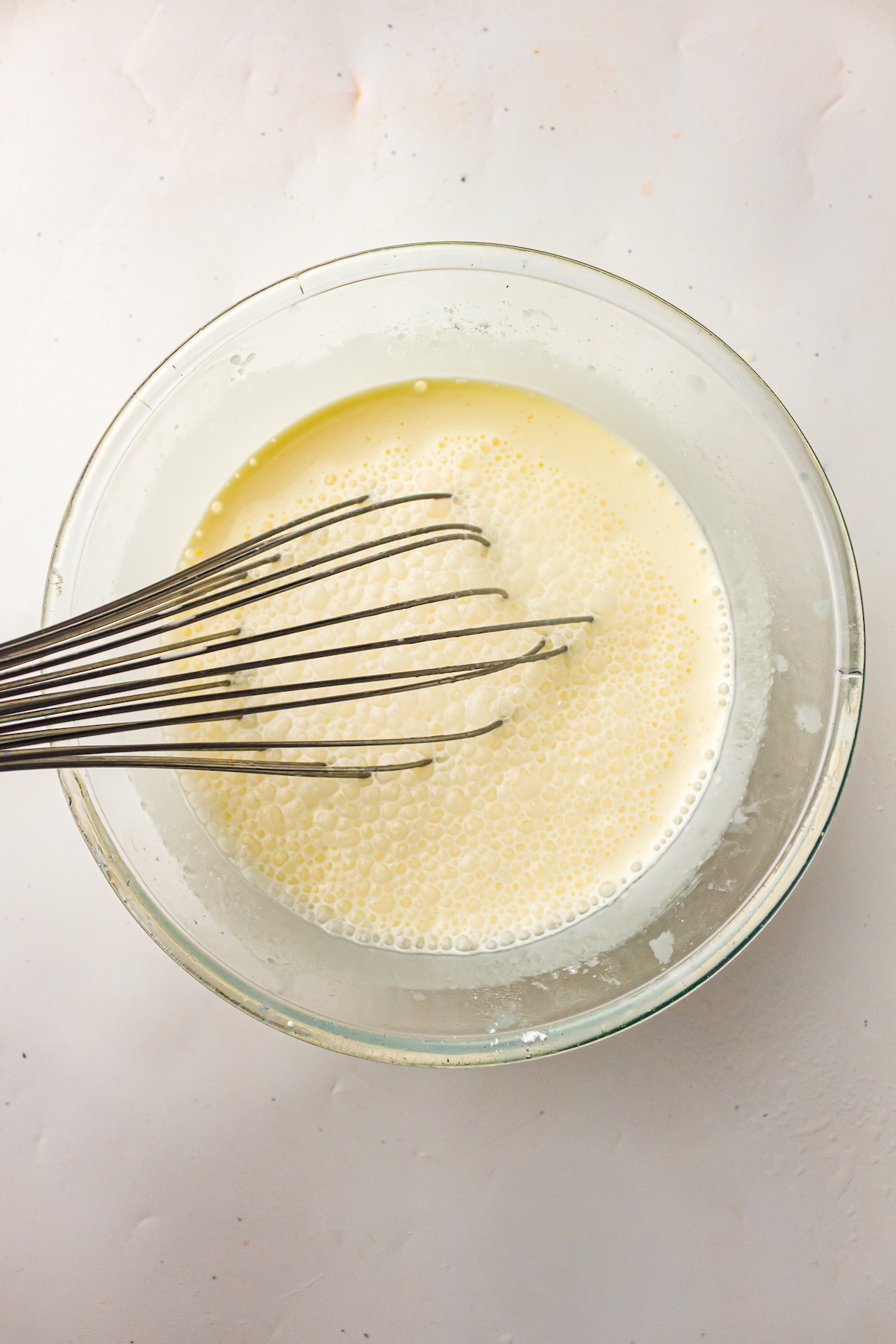 Top-down view of a clear glass bowl with a pale yellow frothy liquid being whisked by a metal whisk, set on a light background.