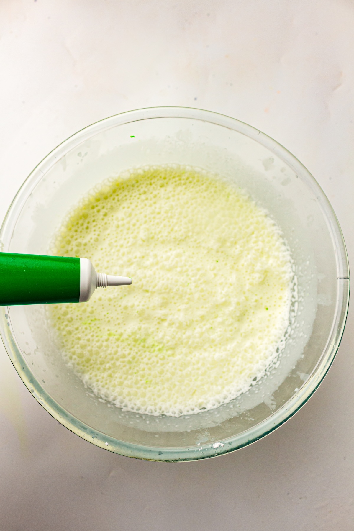 Overhead shot of a clear glass bowl containing a light yellow-green frothy liquid, with a green food coloring tube held above the bowl, set on a neutral background.