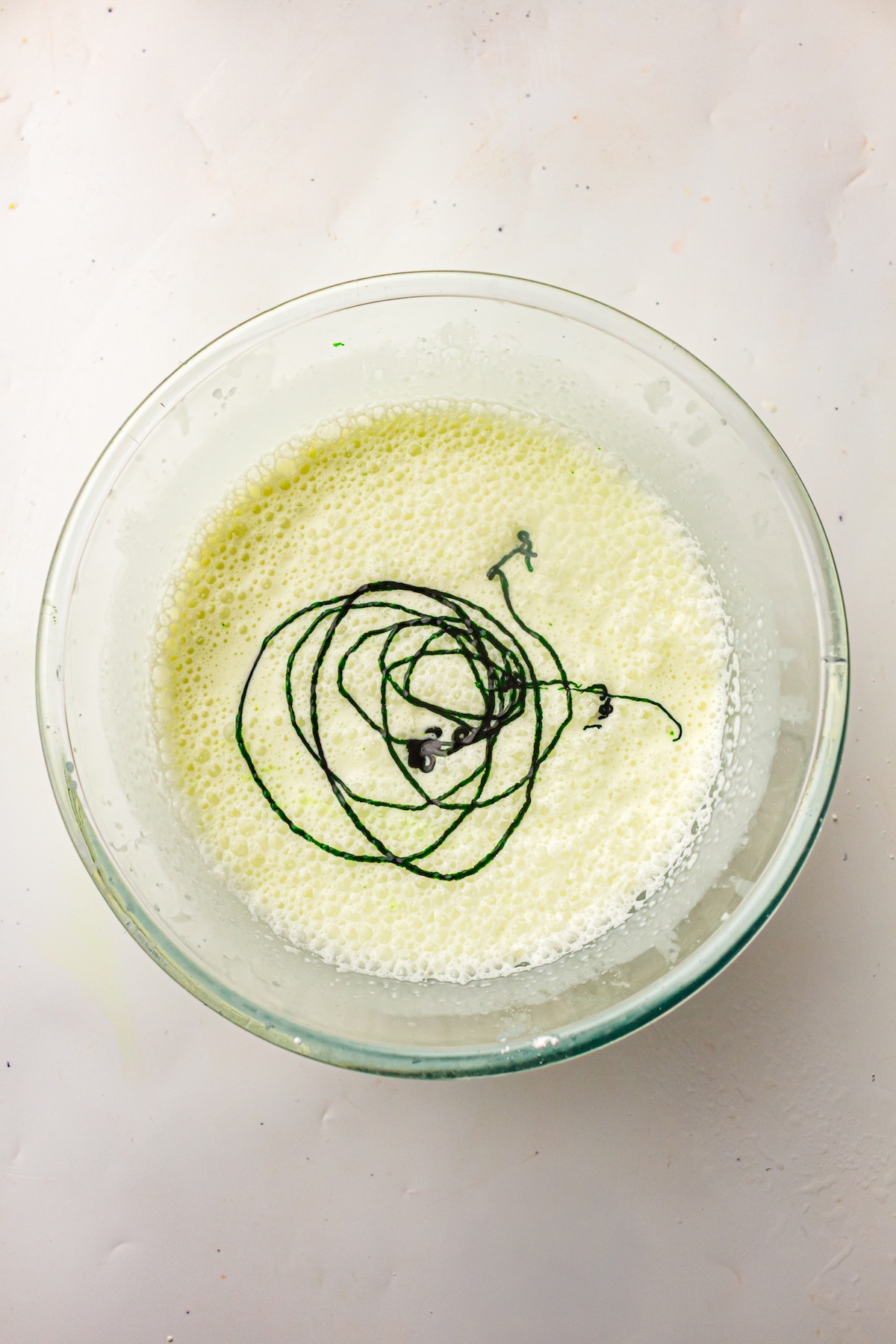 Close-up of a clear glass bowl filled with a light yellow frothy liquid, with dark green food coloring drizzled in swirling lines on the surface, set on a light textured background.