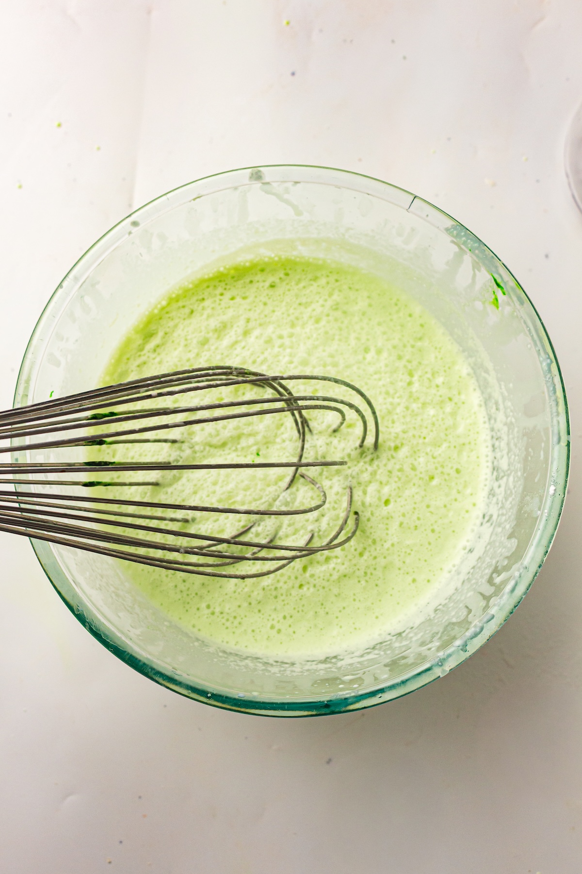 Overhead view of a clear glass bowl containing a light green frothy liquid being actively whisked by a metal whisk, set on a plain light-colored surface.