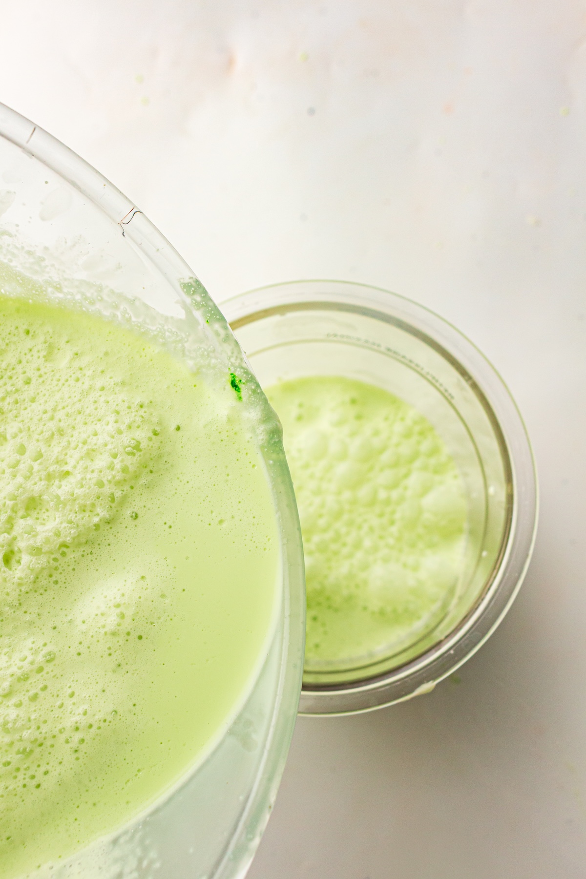 Close-up of a light green frothy liquid being poured from a large clear glass bowl into a smaller clear glass bowl, set on a light background.