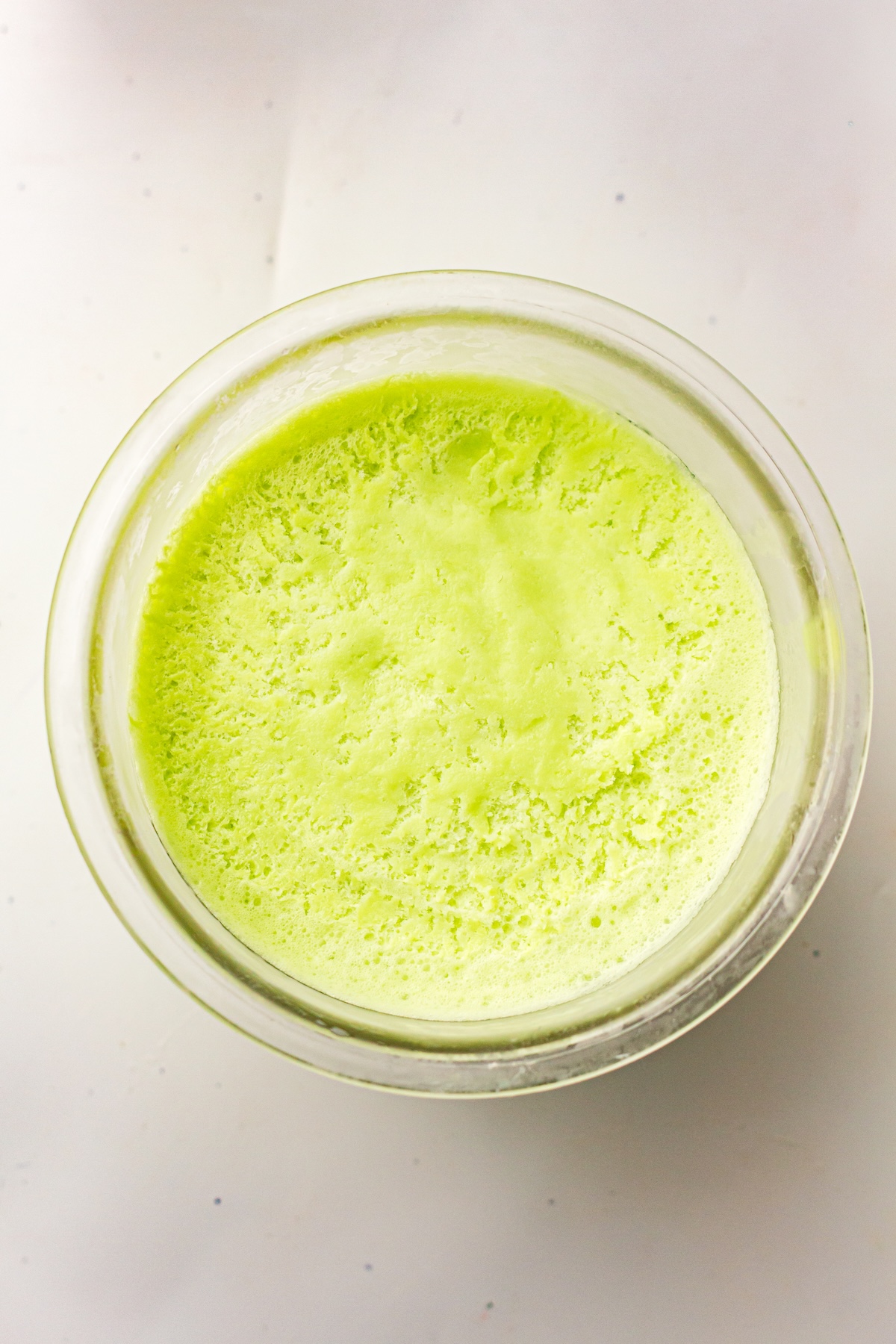 Top-down view of a clear glass bowl filled with a vibrant light green frothy liquid, with a textured surface of small bubbles, set on a light neutral background.