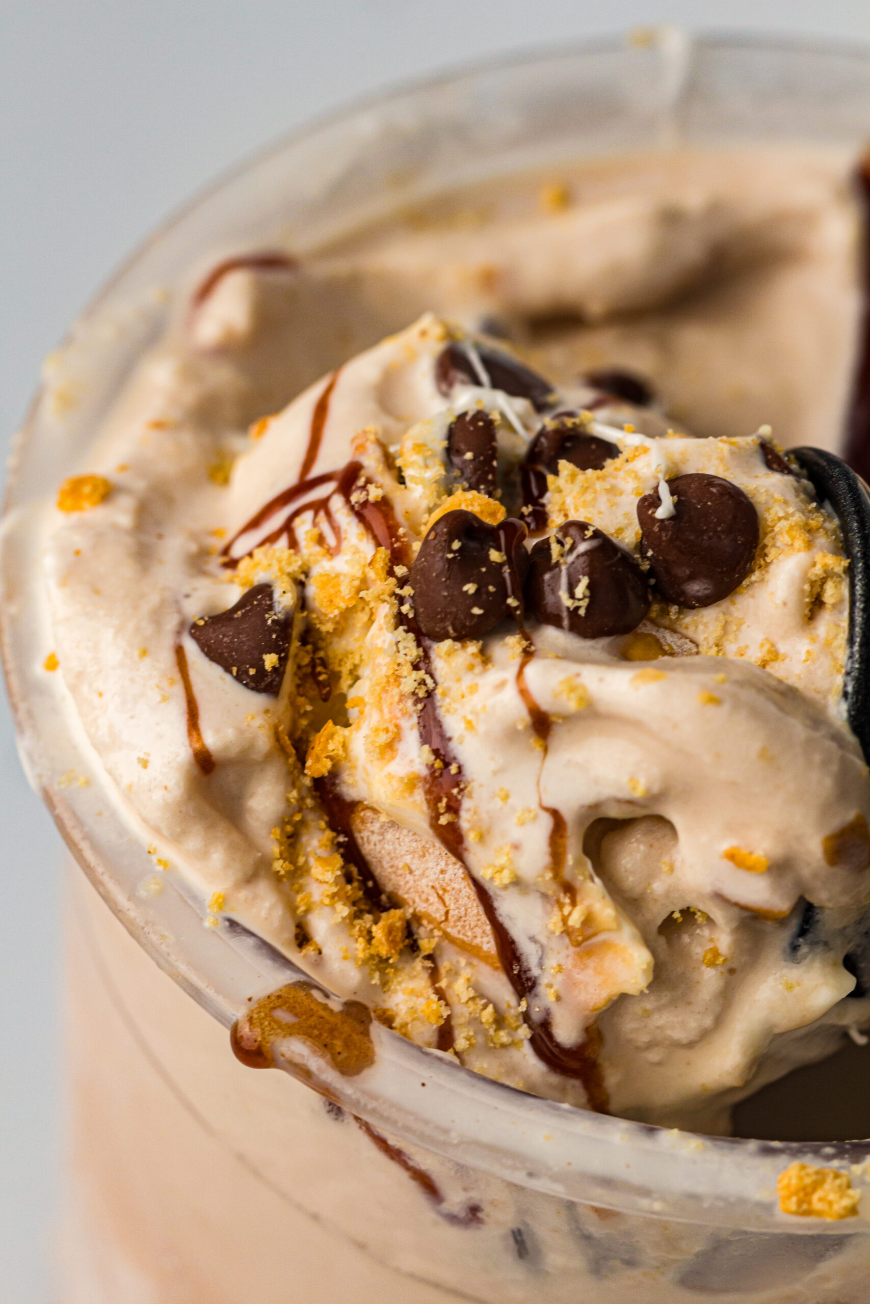 Close-up of light brown ice cream in a clear container, topped with chocolate chips, golden crumbs, and a drizzle of dark syrup, with a dark spoon partially visible on the right.