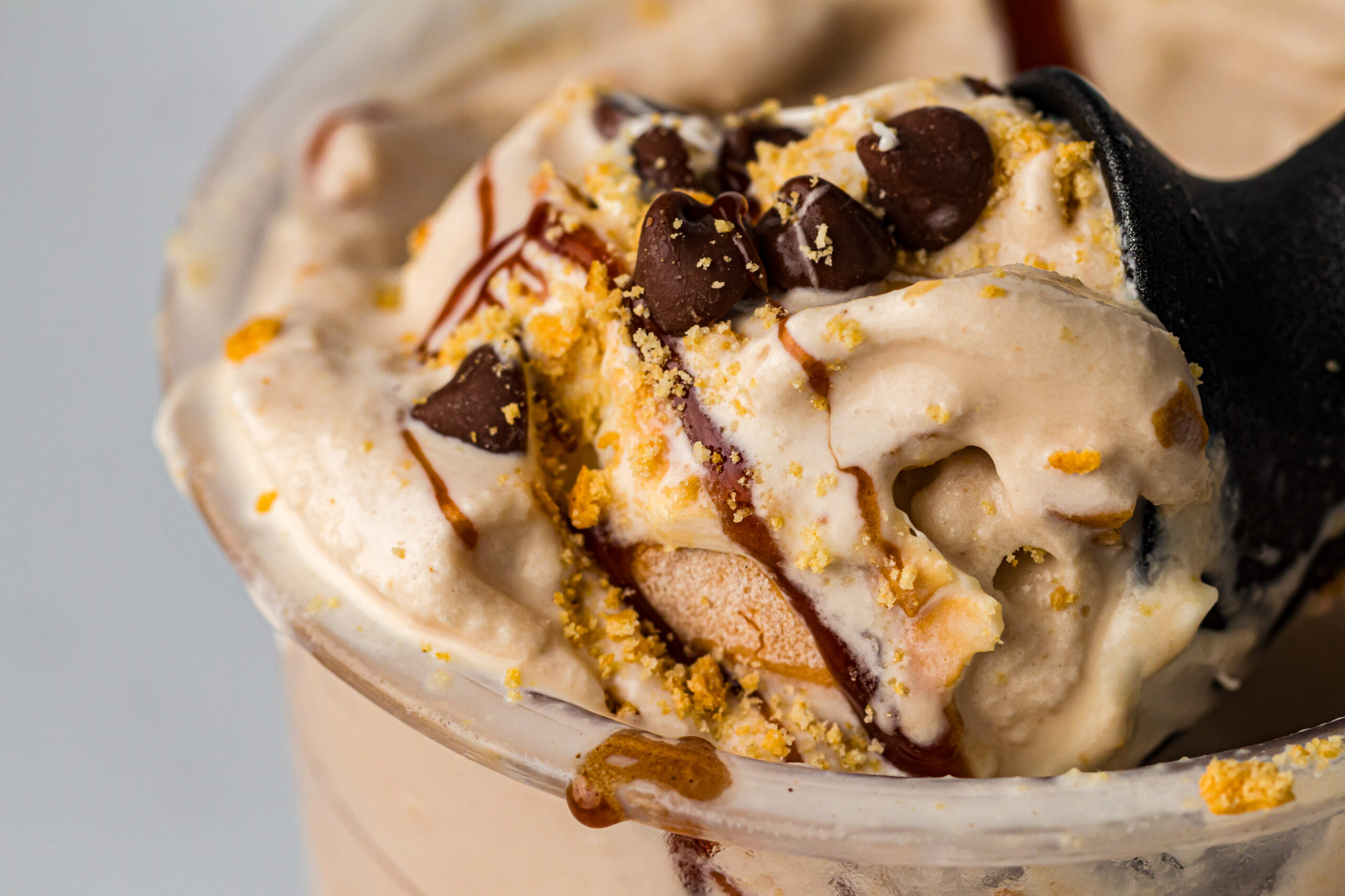 High-angle close-up of creamy light beige ice cream in a clear plastic cup, topped with chocolate chips, golden crumbs, and a drizzle of dark syrup, with a dark spoon on the right.