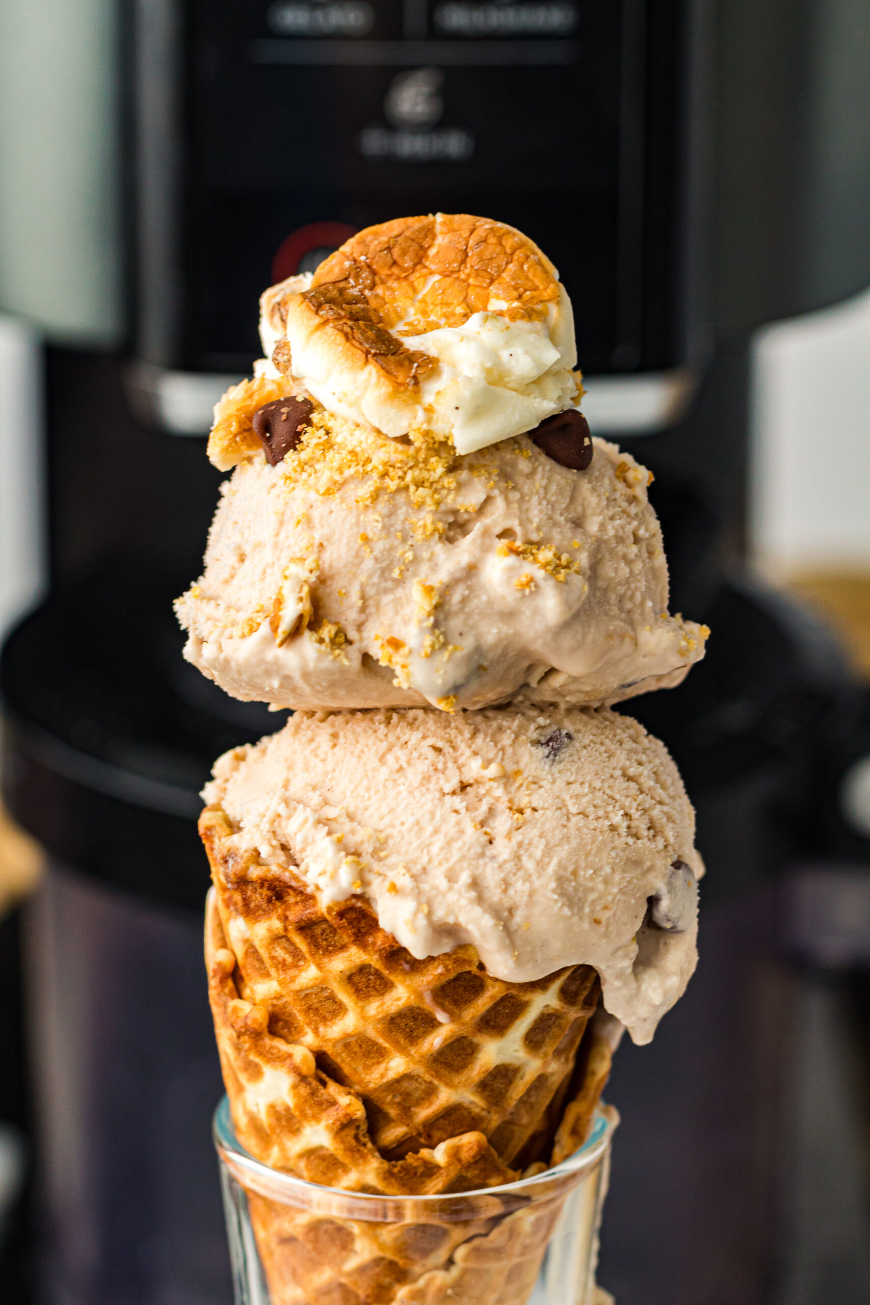 Close-up of a waffle cone with two scoops of light brown ice cream, topped with toasted marshmallow, graham cracker crumbs, and chocolate chips, held in a clear glass against a dark blurred background.
