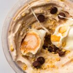 Close-up of light brown ice cream in a clear container, topped with chocolate chips, graham cracker crumbs, and toasted marshmallow pieces, with a clean white background.