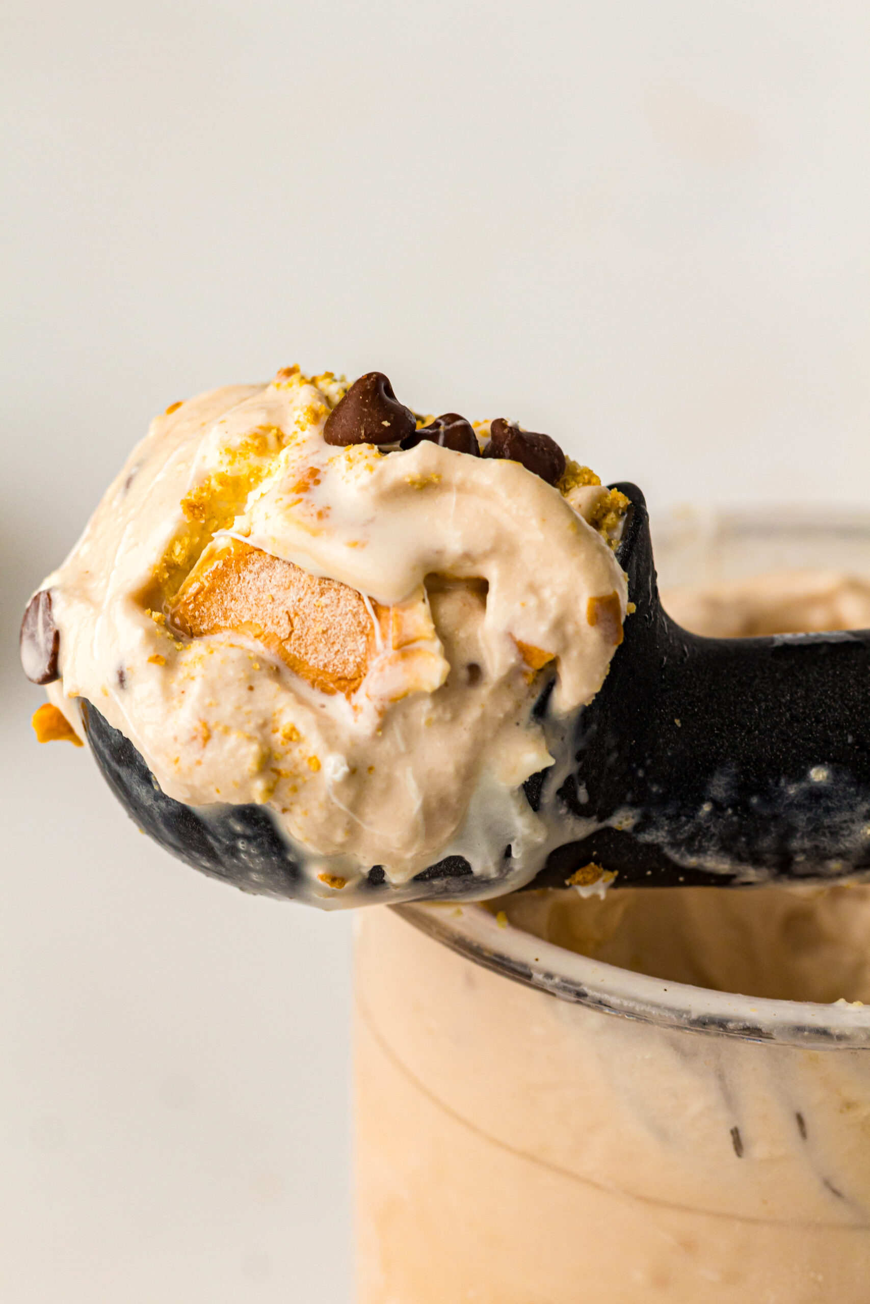 Close-up of a scoop of light brown ice cream with chocolate chips, cookie pieces, and golden crumbs, held in a black ice cream scoop above a clear container.