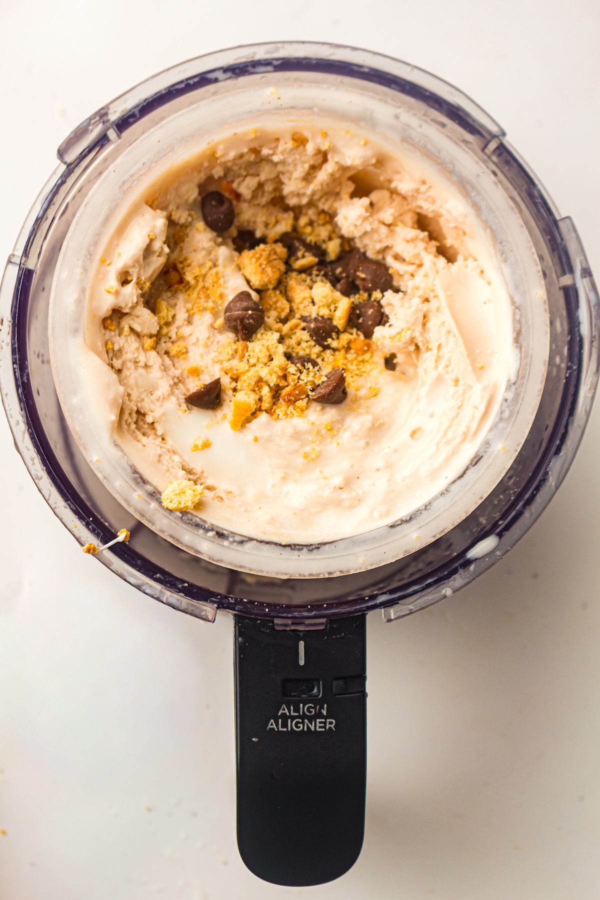Overhead view of a clear container from a kitchen appliance, filled with light tan creamy ice cream, topped with dark chocolate chips and lighter brown crumbs, with a black handle labeled 'ALIGN ALIGNER,' set on a white background.