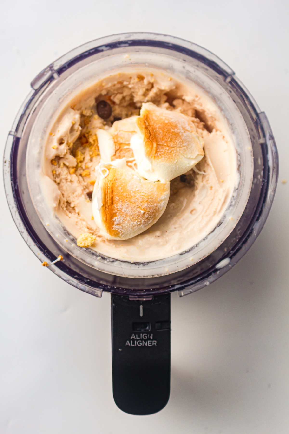 Top-down view of a clear container holding light brown creamy ice cream topped with toasted marshmallows, chocolate chips, and crumbs, with a black appliance handle labeled 'ALIGN ALIGNER,' set on a light countertop.